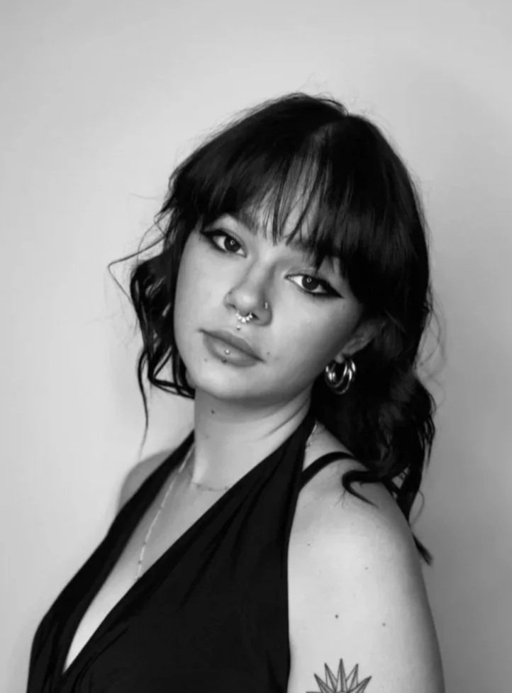 Black and white portrait of a young woman with wavy hair, dark eyeliner, and multiple facial piercings, wearing a sleeveless top and hoop earrings.