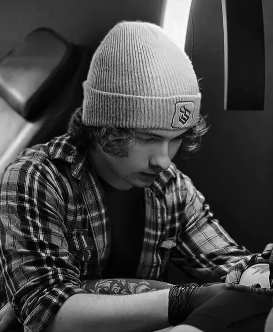 A young man with curly hair, wearing a beanie and plaid shirt, sitting and looking at his phone in black and white.