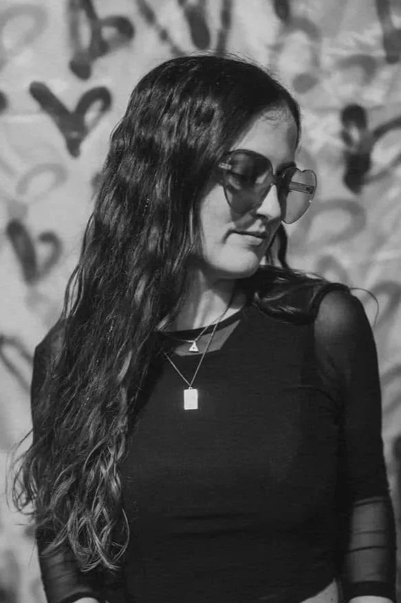 A young woman with long wavy hair wearing sunglasses, a black top, and layered necklaces, looking down with a graffiti-style wall in the background.