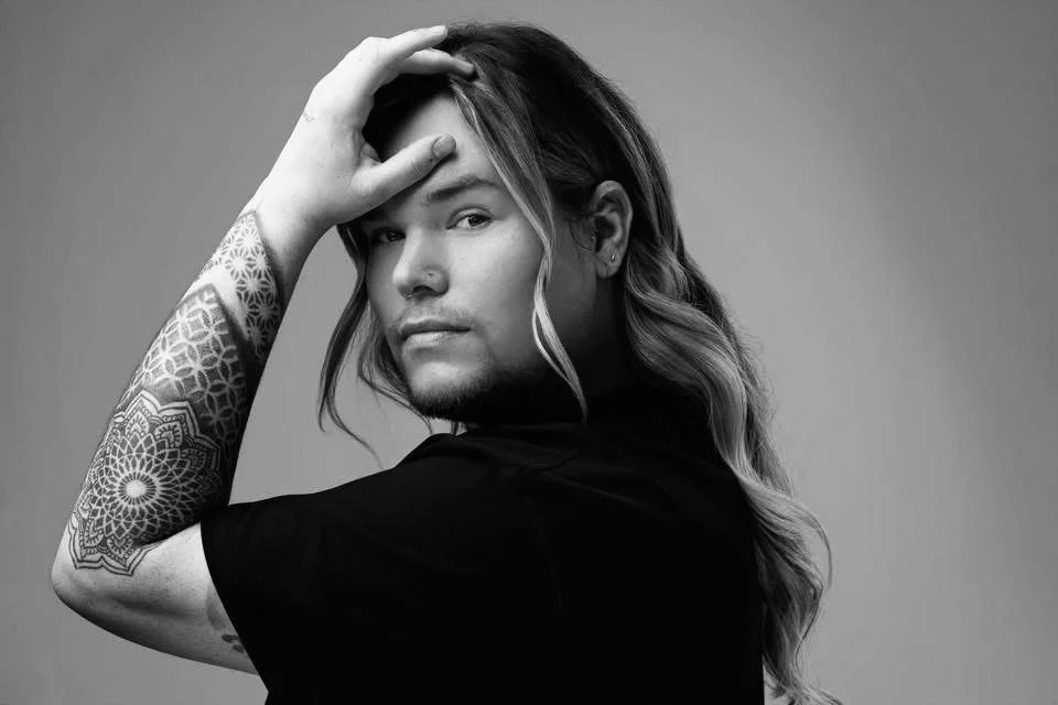 A black and white portrait of a young person with long wavy hair, tattoos on their arm, wearing a black shirt, looking at the camera with one hand on their forehead.
