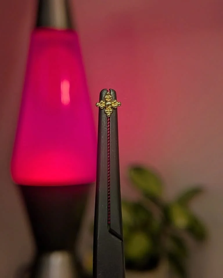 A close-up view of a pair of tweezers holding a gold bee-shaped ring. In the background, there is a pink and black lava lamp and some blurred green leaves.