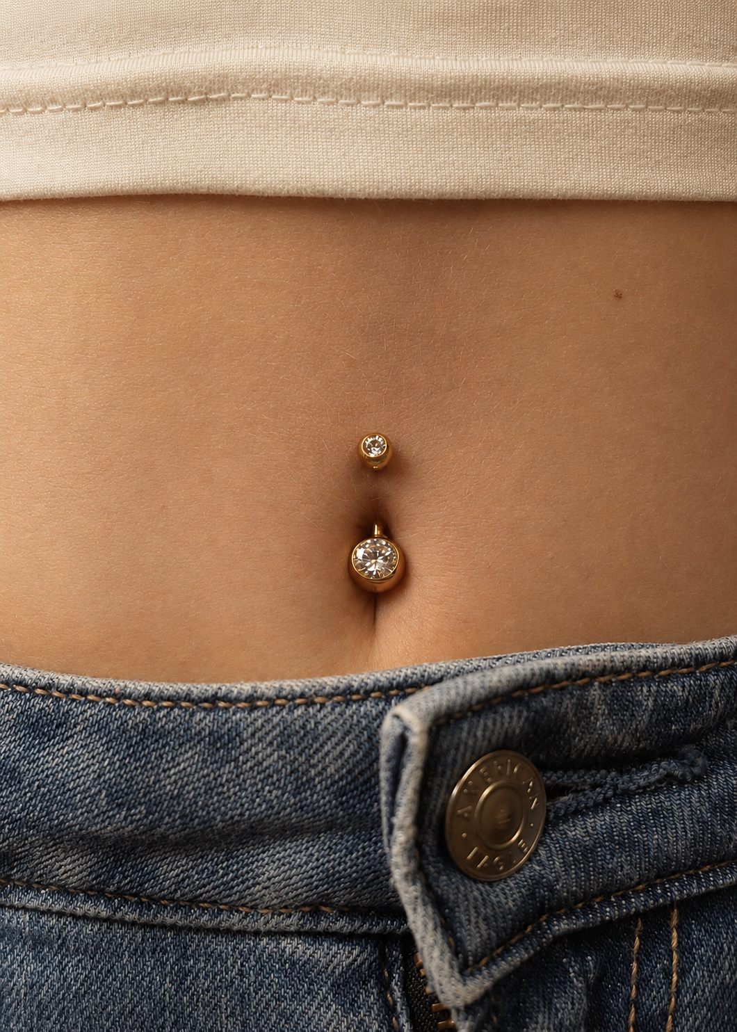 Close-up of a woman's bare midriff with a belly button piercing featuring two small, round, sparkling gemstones. She is wearing a white top and blue jeans.
