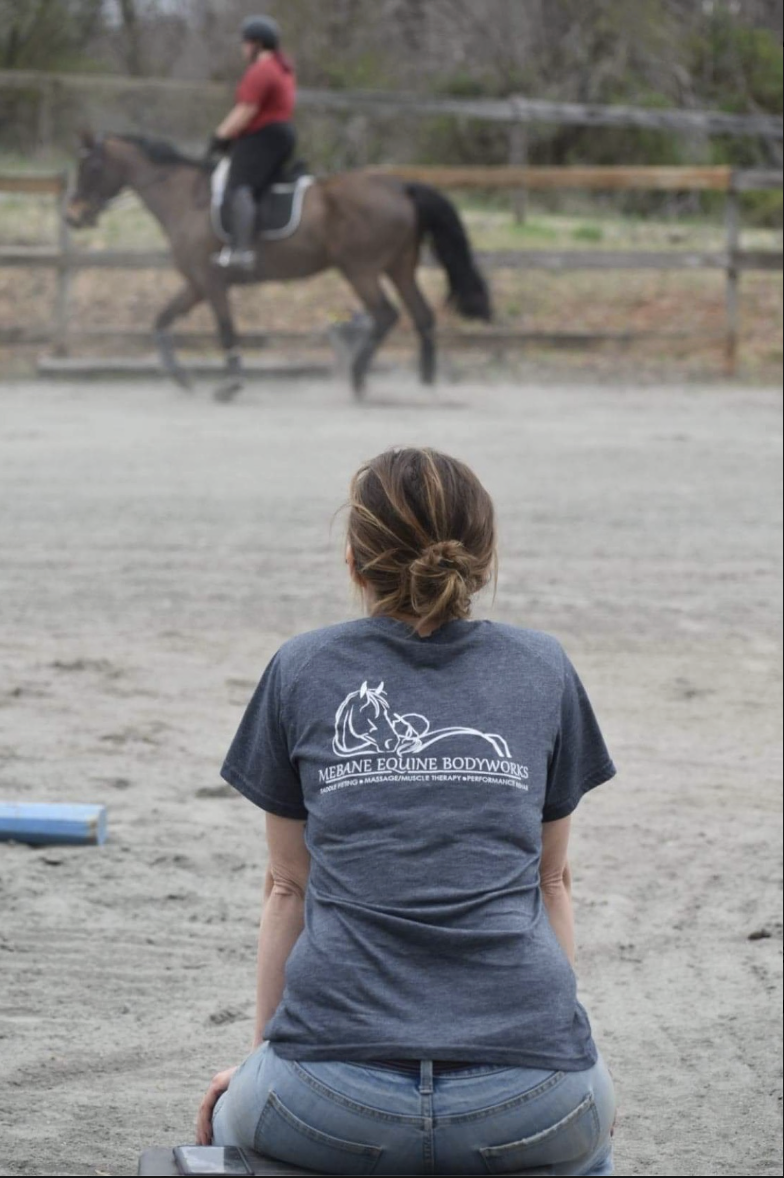 Equestrian consultant Tara Mebane wearing a Mebane Equine Bodyworks shirt while observing a sport horse and rider to evaluate movement patterns and saddle balance.