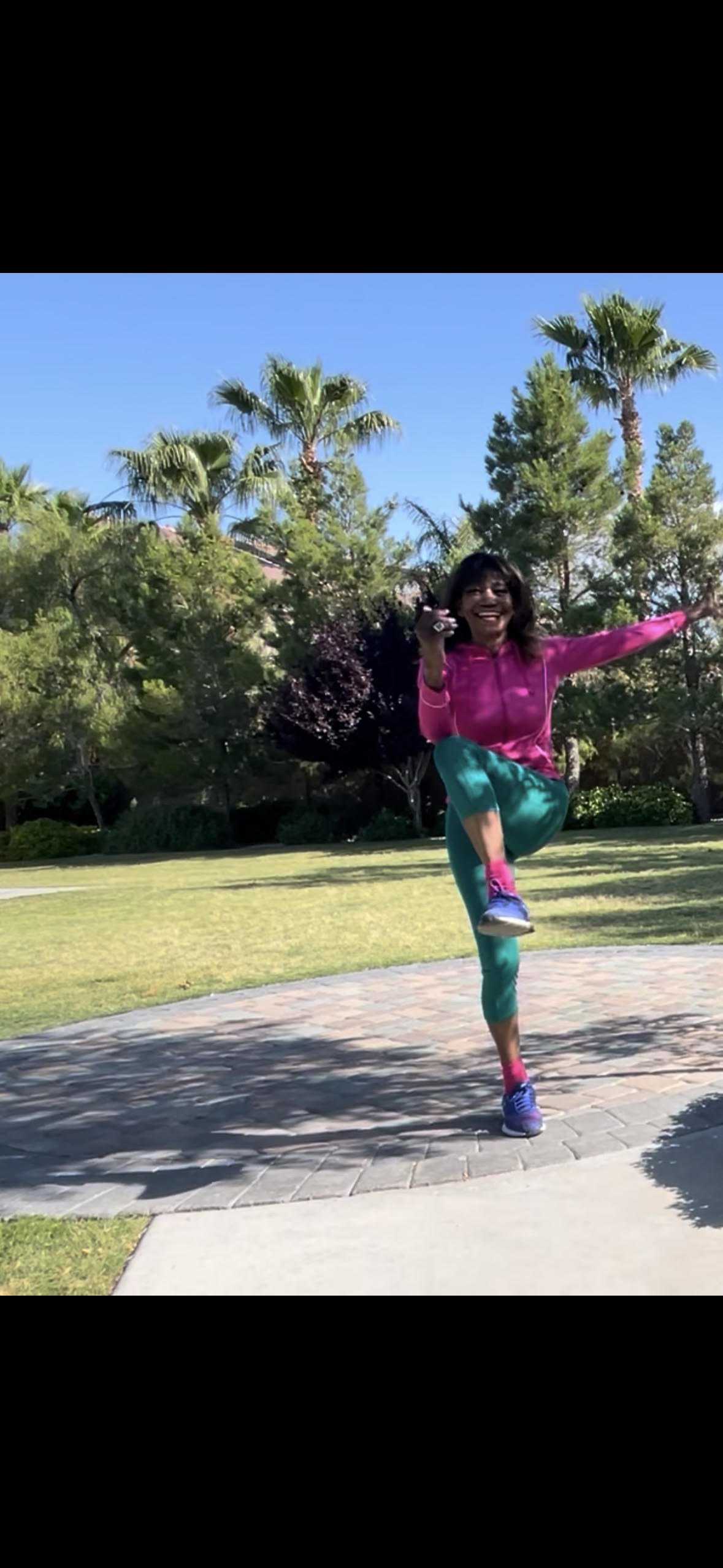 A woman in colorful athletic clothing, including a pink jacket, teal leggings, pink socks, and purple running shoes, is outdoors on a sunny day. She is balancing on one foot with her knee lifted, arms outstretched, smiling with trees and blue sky in the background.