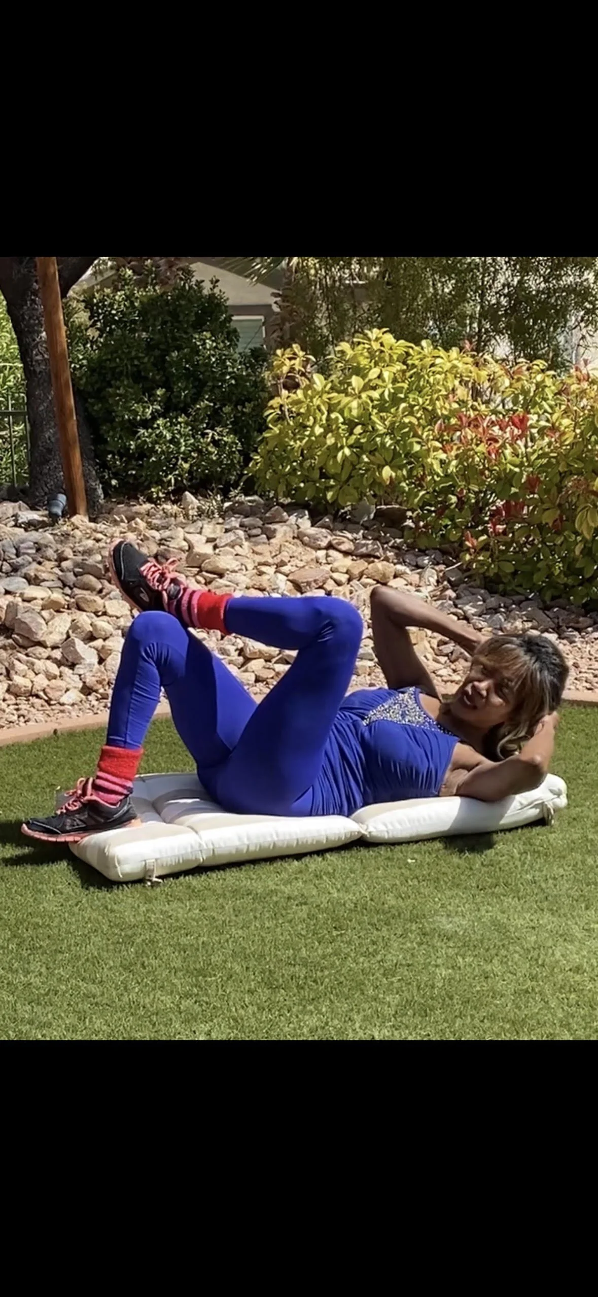 Woman lying on her side on an outdoor mattress, performing a leg exercise with her knee bent and foot raised, in a garden with shrubs and rocks in the background.