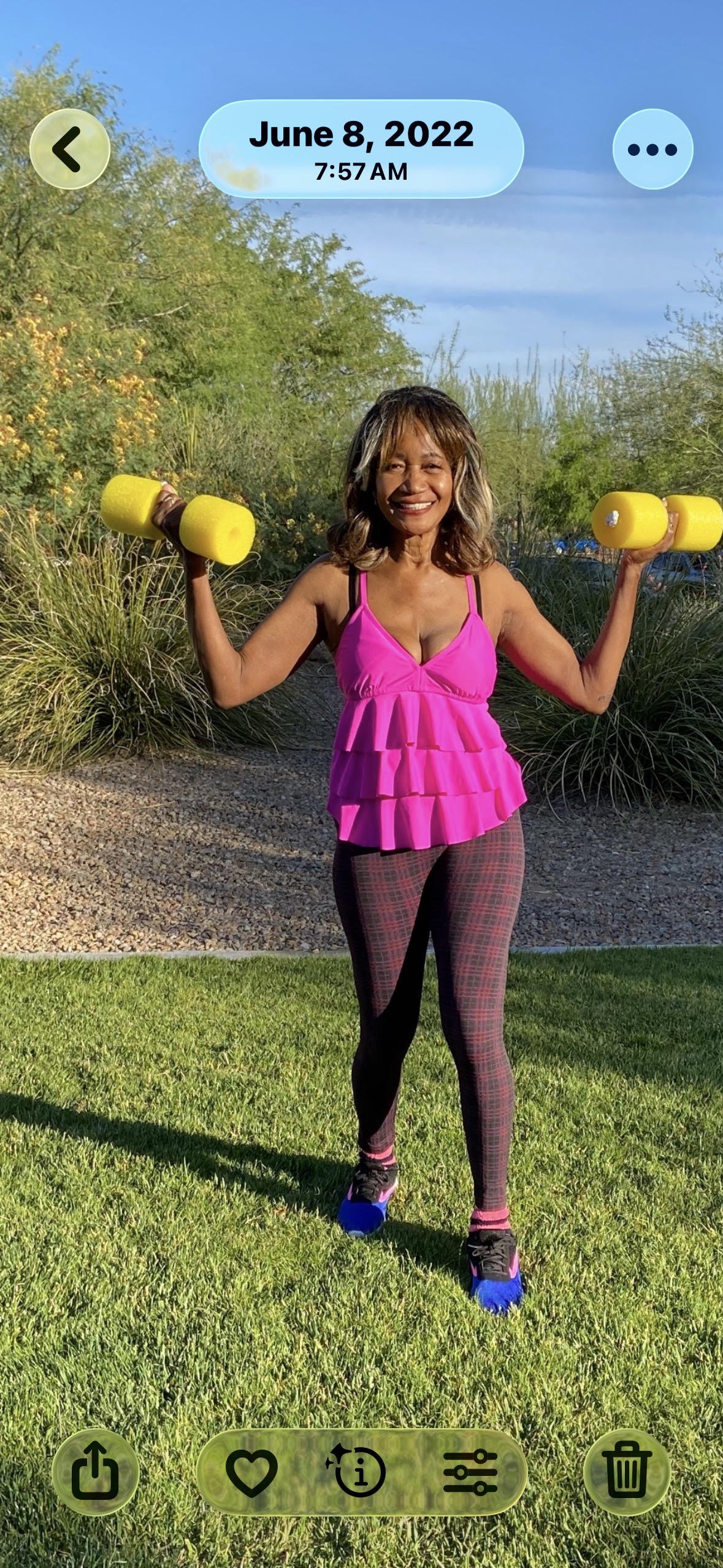 A woman in a bright pink top and patterned leggings holding yellow dumbbells outdoors on a sunny day.