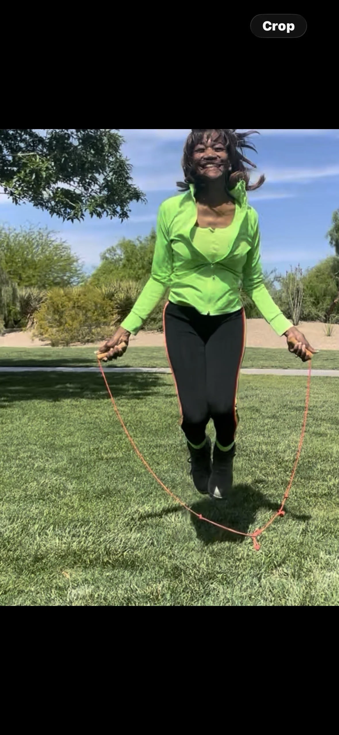 Woman skipping rope on a grassy park with trees and a blue sky.