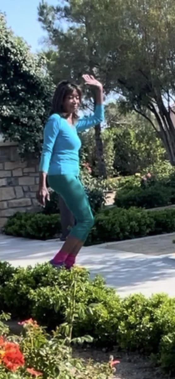 A woman wearing a bright blue long-sleeve shirt and teal capri leggings with pink socks is standing outdoors on a sunny day, surrounded by green bushes and trees, with her right hand raised as if waving or shielding her eyes from the sun.