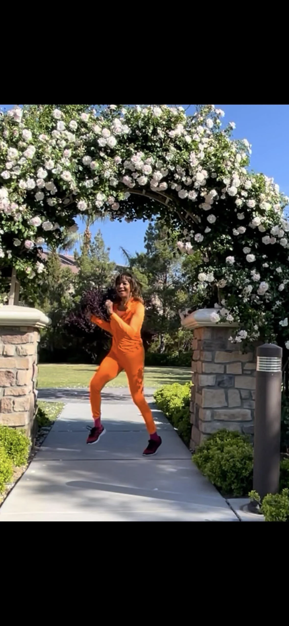 A woman jumping on a sidewalk under a floral archway in a residential neighborhood, wearing an orange outfit and black shoes.