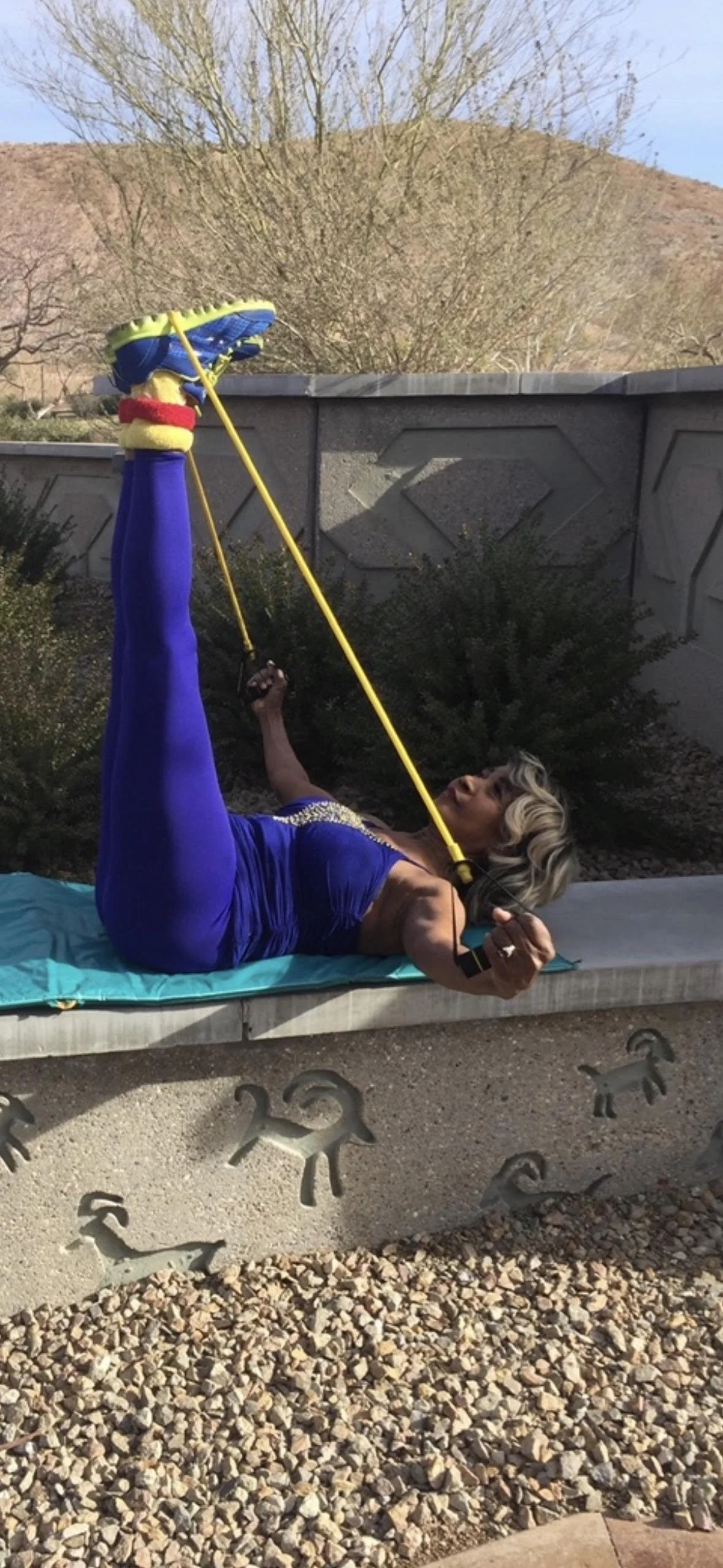 A woman in a blue dress lying on a bench outdoors, holding a yellow exercise stretch band with her foot elevated and hooked through it.