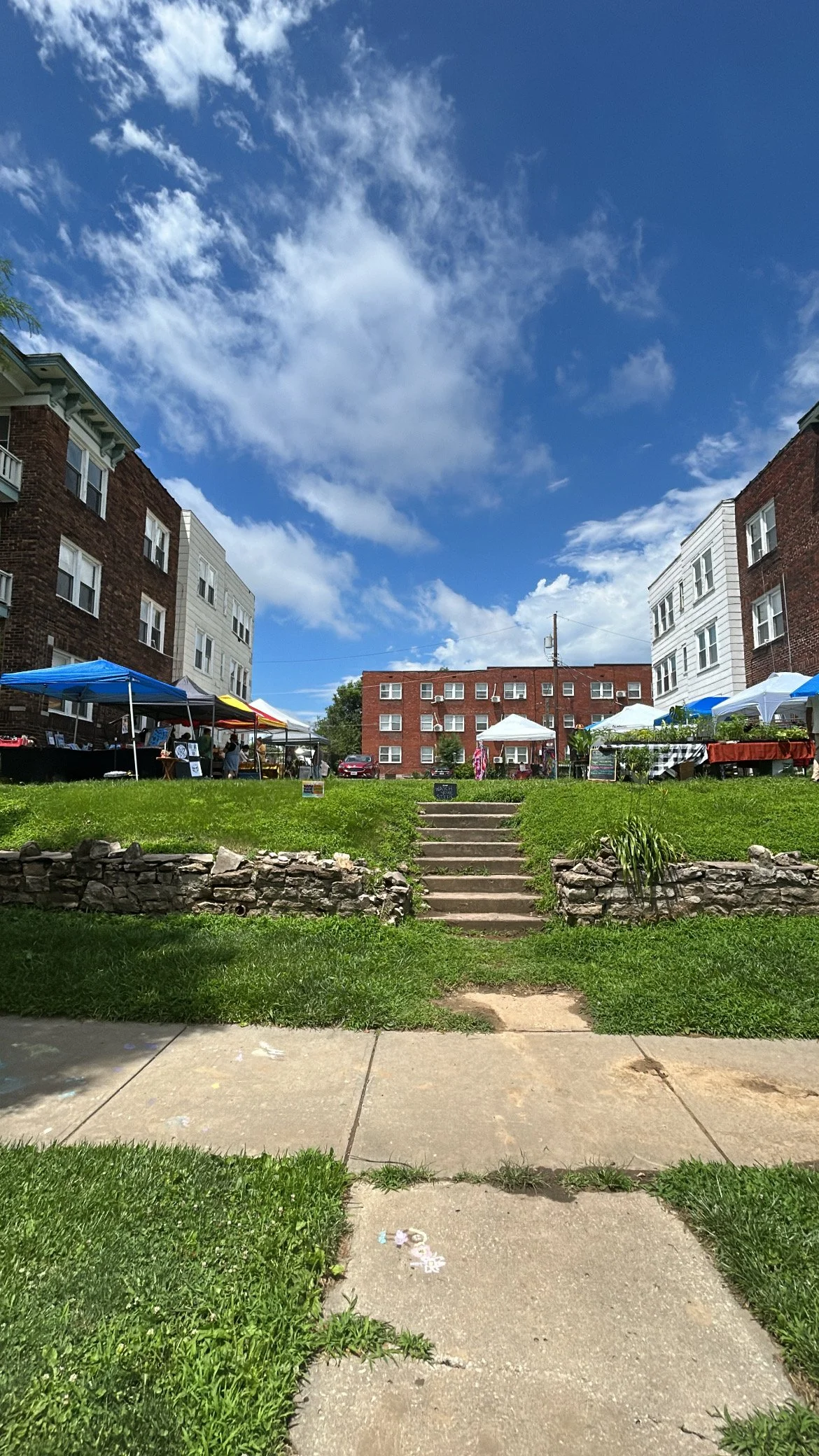 View of an outdoor marketplace set up on a grassy area between residential buildings on a bright, partly cloudy day. Tents and tables with vendors' goods are visible, with steps leading up to the market area from the sidewalk.