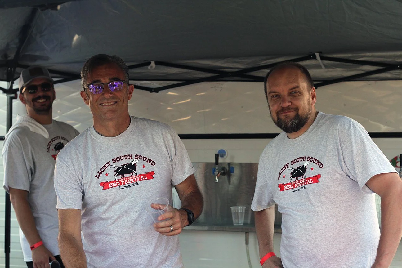 Three men wearing matching gray t-shirts with a barbecue festival logo, standing under a canopy at an outdoor event.
