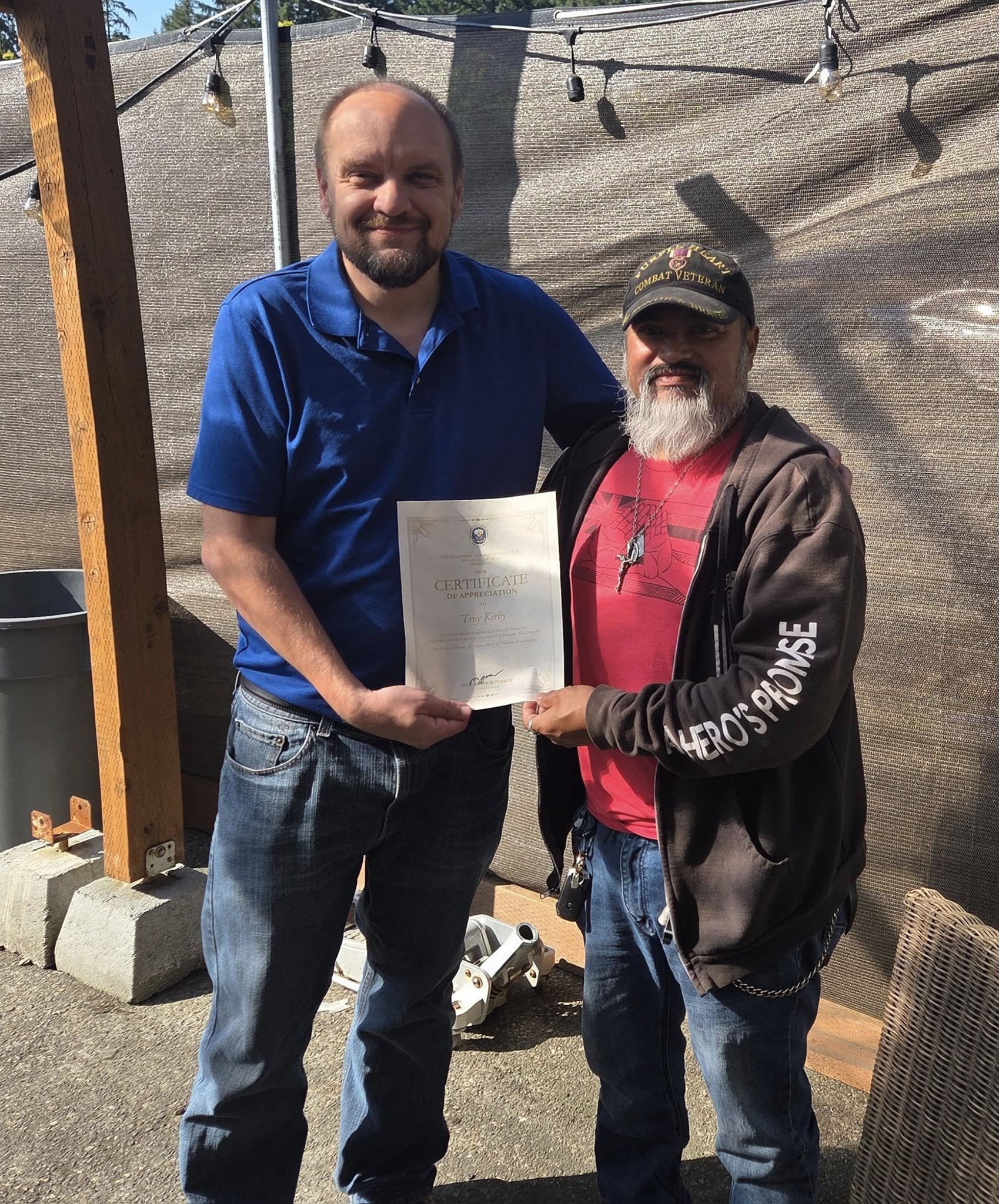 Two men standing outdoors, one holding a certificate of appreciation, posing for a photo. The man on the left has a beard, glasses, and is wearing a blue polo shirt. The man on the right has a beard, is wearing a black cap, pink t-shirt, black jacket, and jeans. The background includes a wooden post, string lights, and outdoor items.