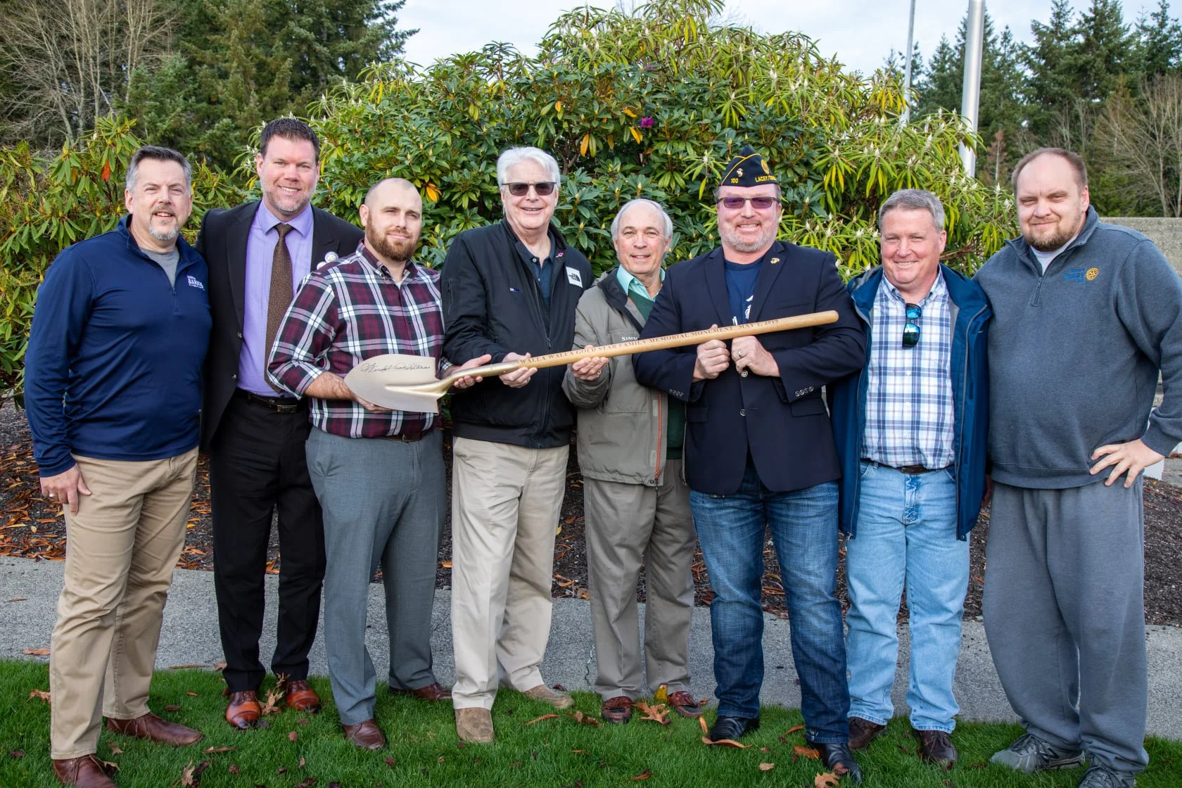 A group of nine men standing outdoors in front of a large green bush, smiling and holding a ceremonial shovel with engraved text, at a dedication event.
