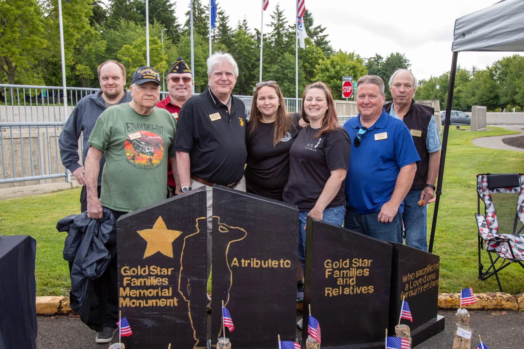 Group of eight people standing outdoors, posing in front of a memorial monument with a black and gold sign that reads 'Gold Star Families Memorial Monument.' Some individuals wear military-related clothing and hats, and small American flags are displayed at the base of the monument.