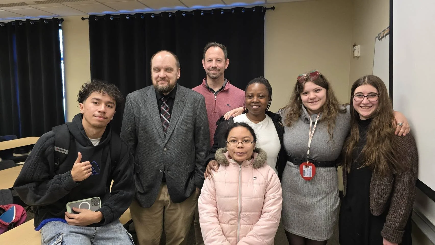 Group of seven diverse people, including students and adults, smiling and posing together in a classroom with black curtains and a whiteboard.