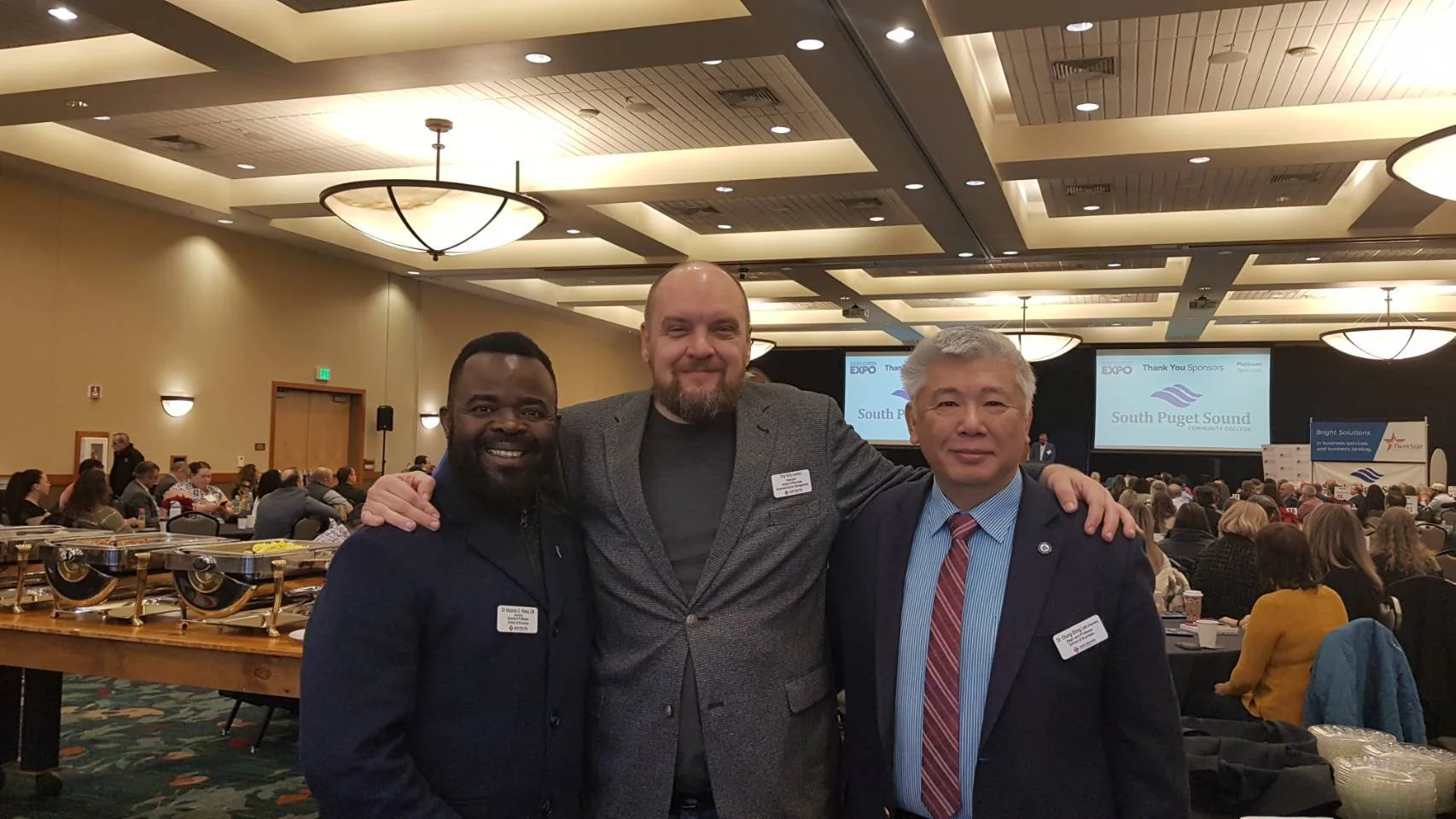 Three men standing together at a conference, smiling for the camera with a large audience and presentation screens in the background.