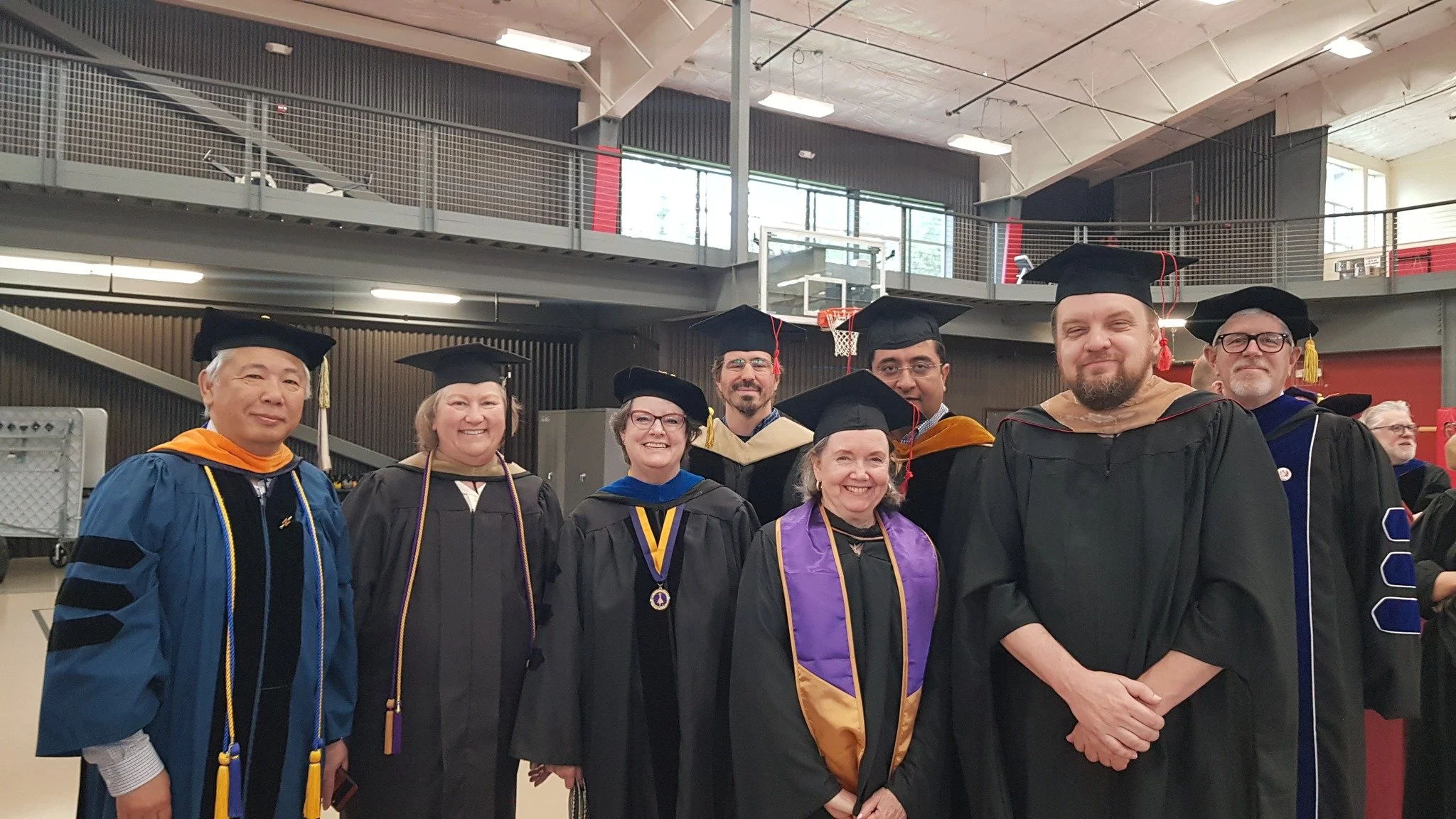 A group of people wearing academic regalia and caps standing together at a graduation ceremony inside a gymnasium or auditorium.