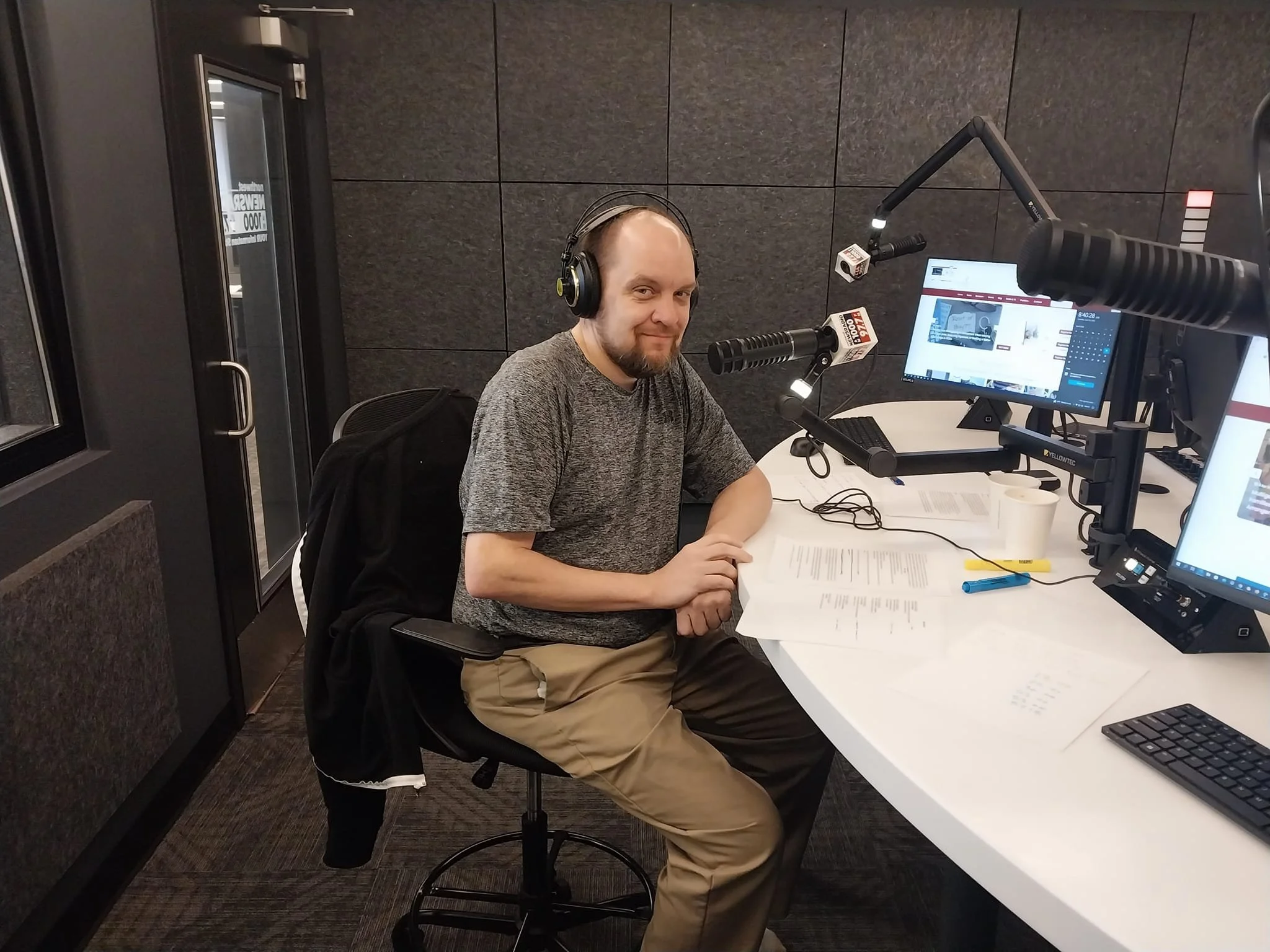 A man sitting at a radio broadcast studio desk, wearing headphones, smiling at the camera, with microphones, computers, papers, and coffee cups on the desk.