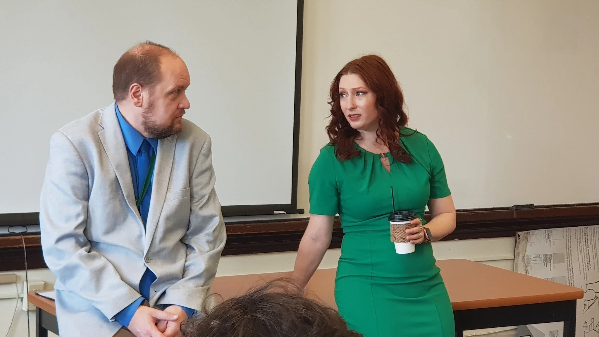 A man and a woman are standing and talking in a classroom or meeting room. The man has a beard, is wearing a light-colored blazer and blue shirt, and looks at the woman. The woman has red hair, is wearing a green dress, and is holding a paper coffee cup.