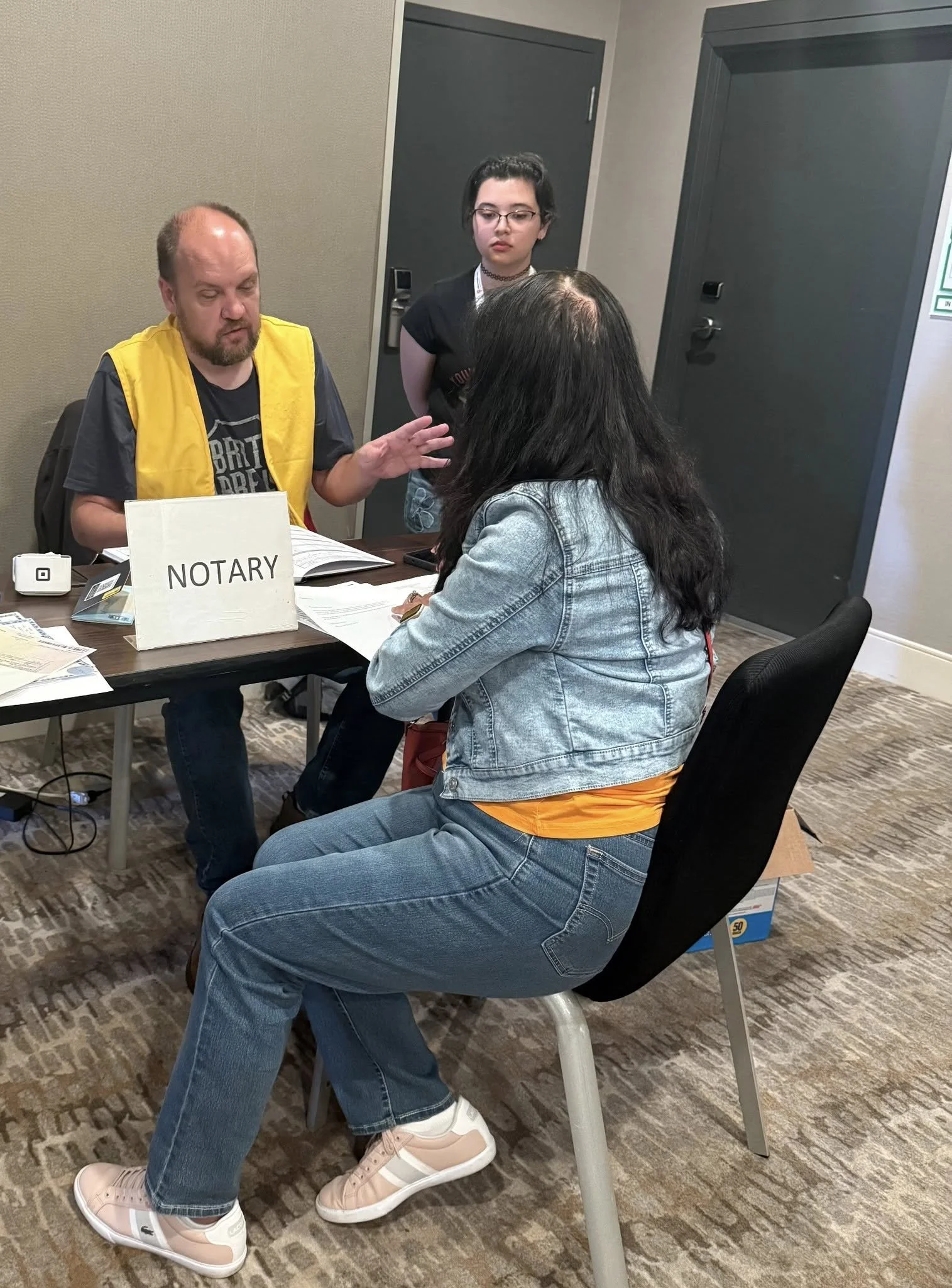 A man wearing a yellow vest and a woman in a denim jacket sitting at a table having a discussion with another woman standing beside them, with a sign that says 'NOTARY' on the table.
