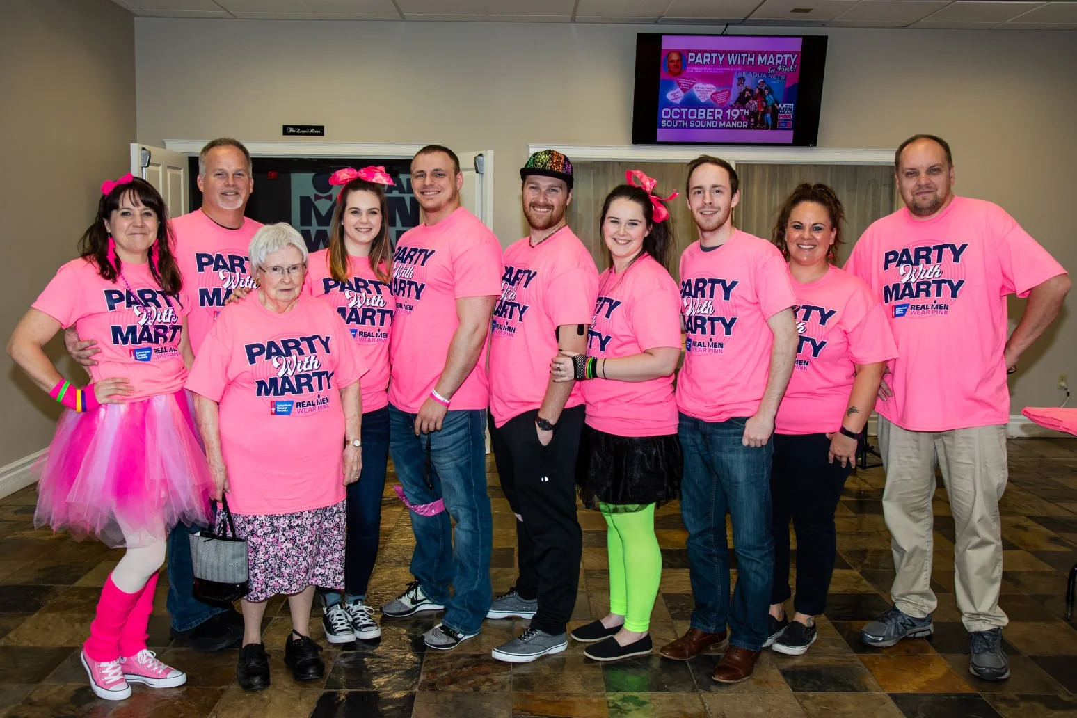 A group of eleven people dressed in pink shirts with a pink tutu and leg warmers, posing indoors in front of a television screen at an event. They are smiling and standing close together.