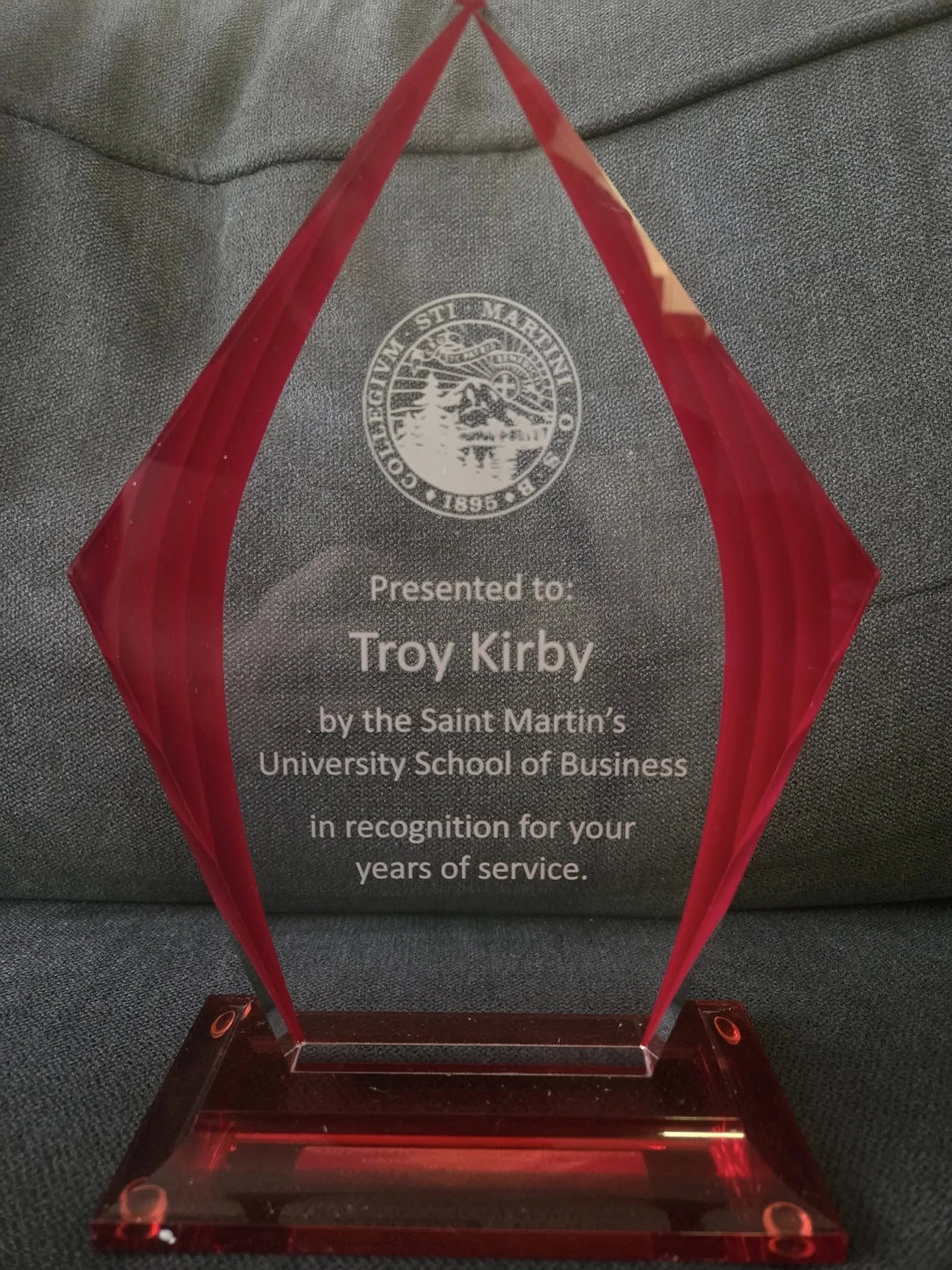 A clear, acrylic award or trophy with a triangular shape, featuring the seal of the University of Saint Martin at the top, and an inscription recognizing Troy Kirby for his years of service.