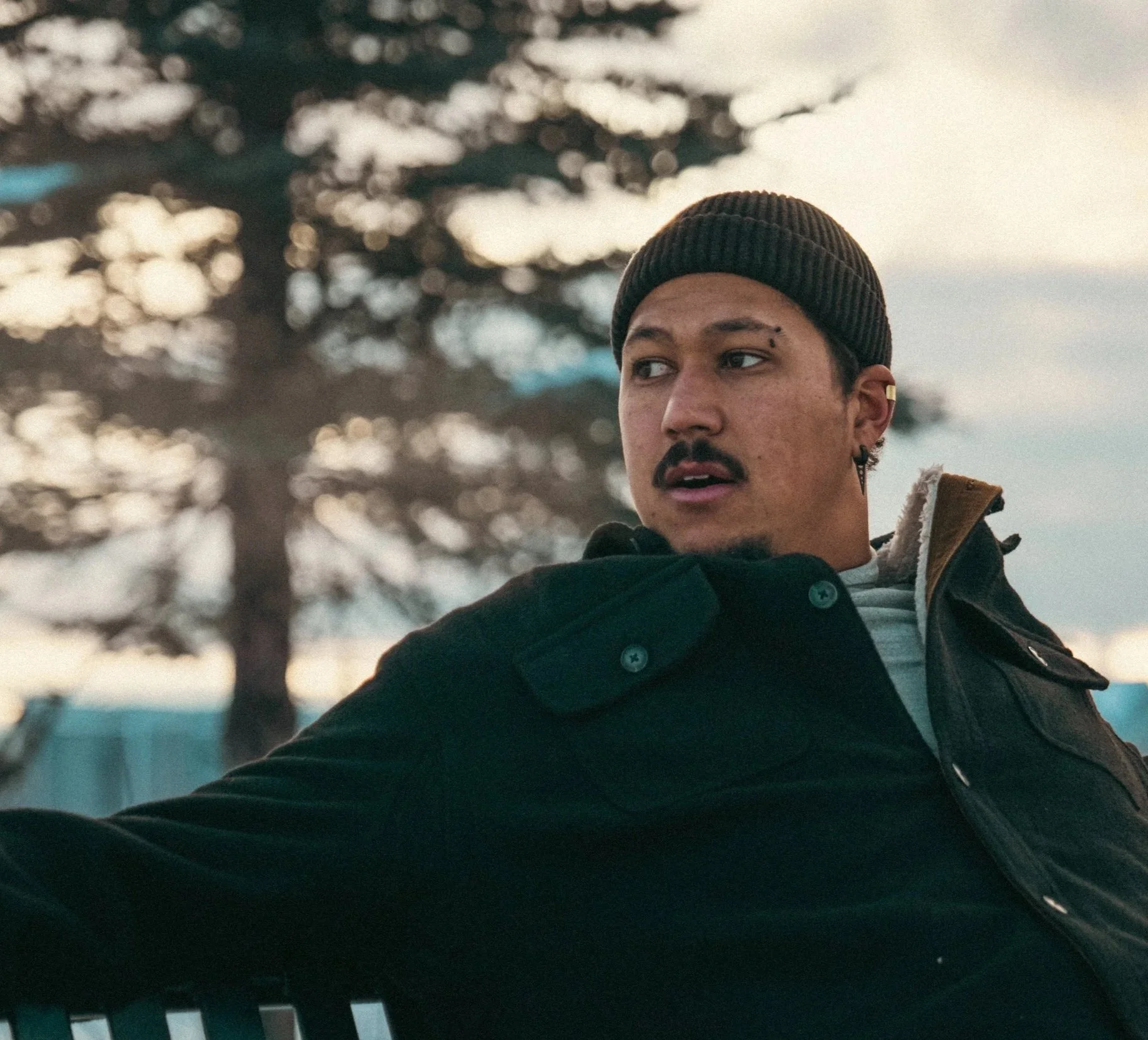 R'Know wearing a brown beanie, earrings, and a black jacket looking to the side outdoors during daytime, with blurred trees and a cloudy sky in the background.