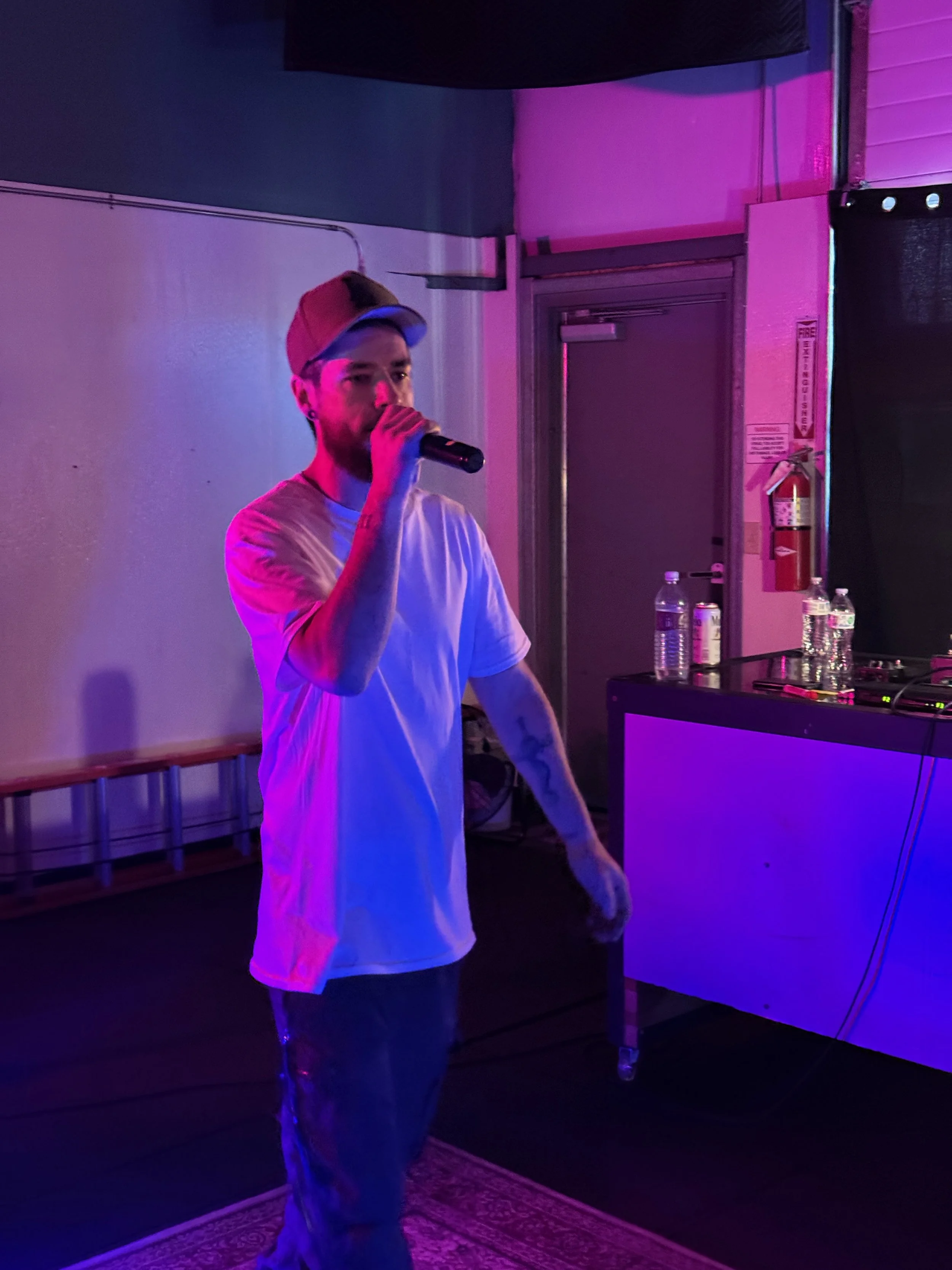 Yung Signal wearing a white t-shirt and baseball cap holding a microphone, standing in a room with pink and purple lighting, near a table with water bottles and cans.