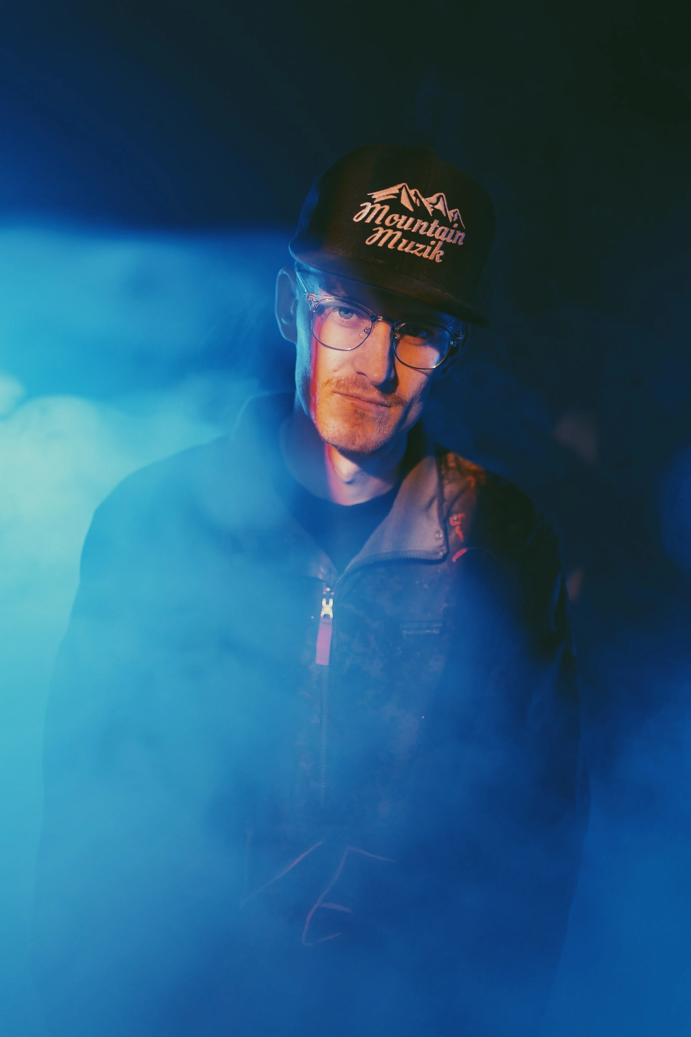TR. wearing a black cap with Mountain Muzik logo, glasses, and a black jacket in a dark setting with blue and teal lighting.