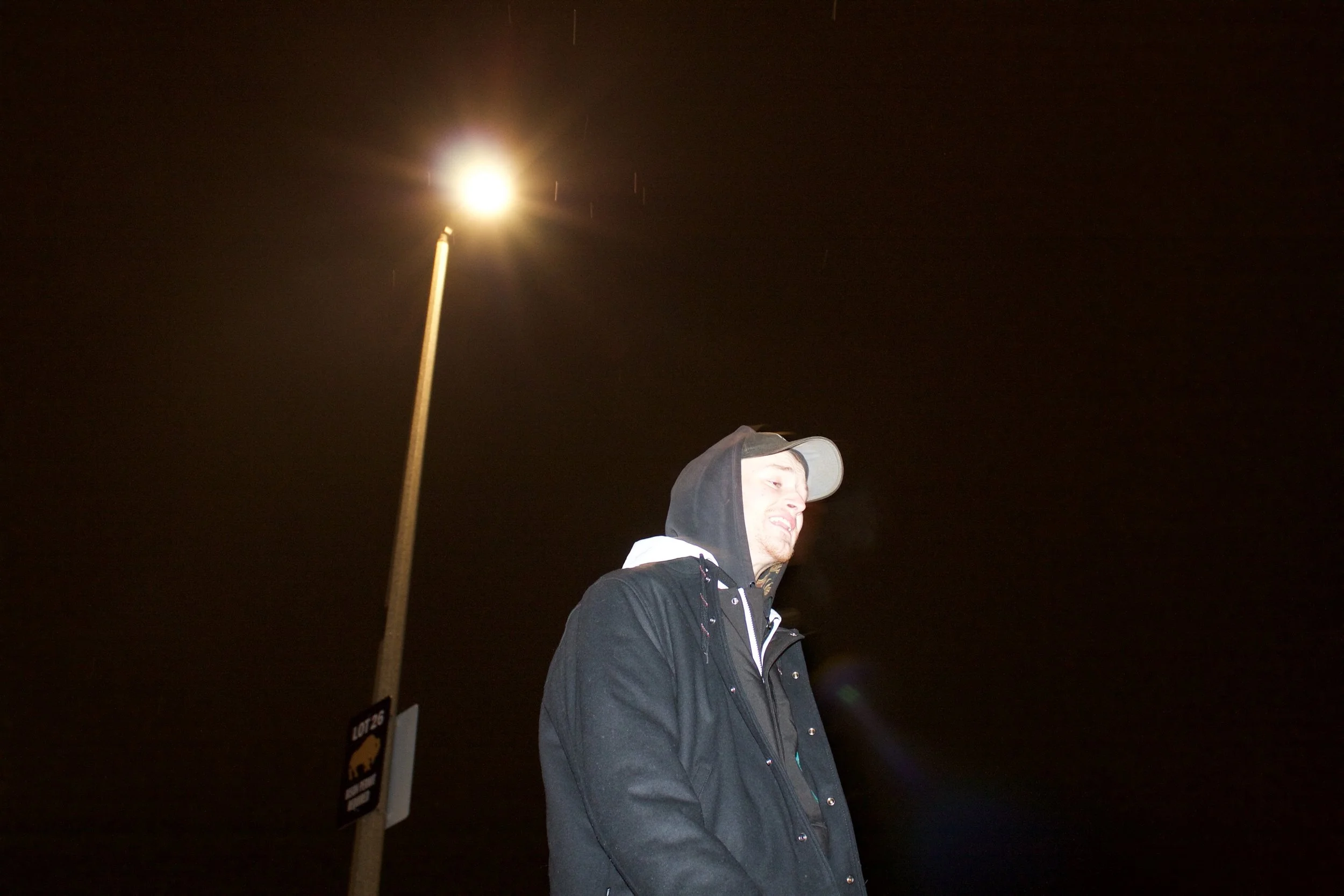 TR. wearing a hoodie and jacket standing outdoors at night, illuminated by a bright streetlight.