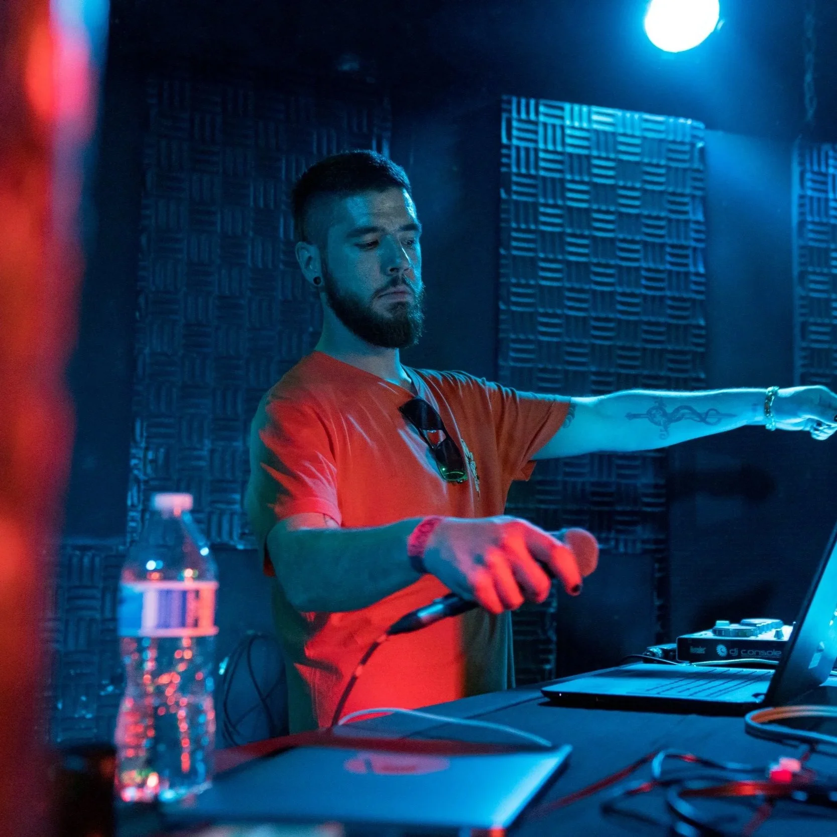 Yung Signal with a beard and earrings is DJing in a dimly lit room with blue lighting. He is wearing a red shirt and has sunglasses hanging from his collar. He is adjusting sound equipment on a table with a laptop, water bottle, and audio gear.
