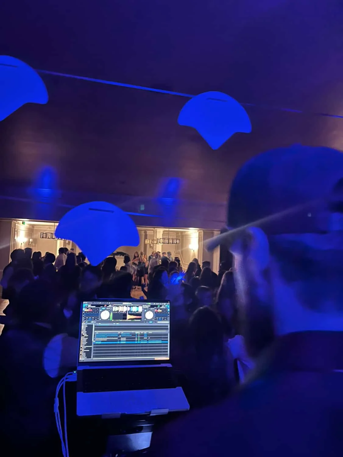 A crowded party or dance event with blue decorative lights hanging from the ceiling, Signal DJing with a laptop mixing music, and people dancing and socializing in a dimly lit room.