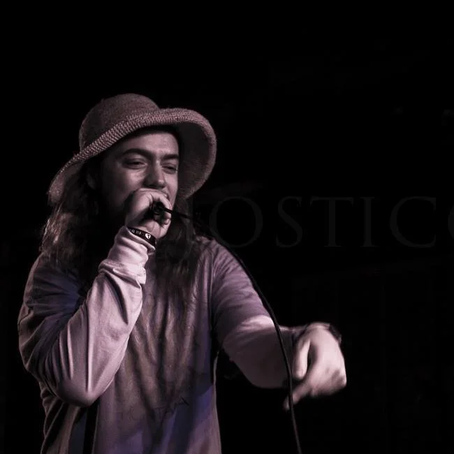 Symon wearing a floppy hat and a long sleeve shirt, smiling and holding a microphone, appears to be performing on stage with a dark background.