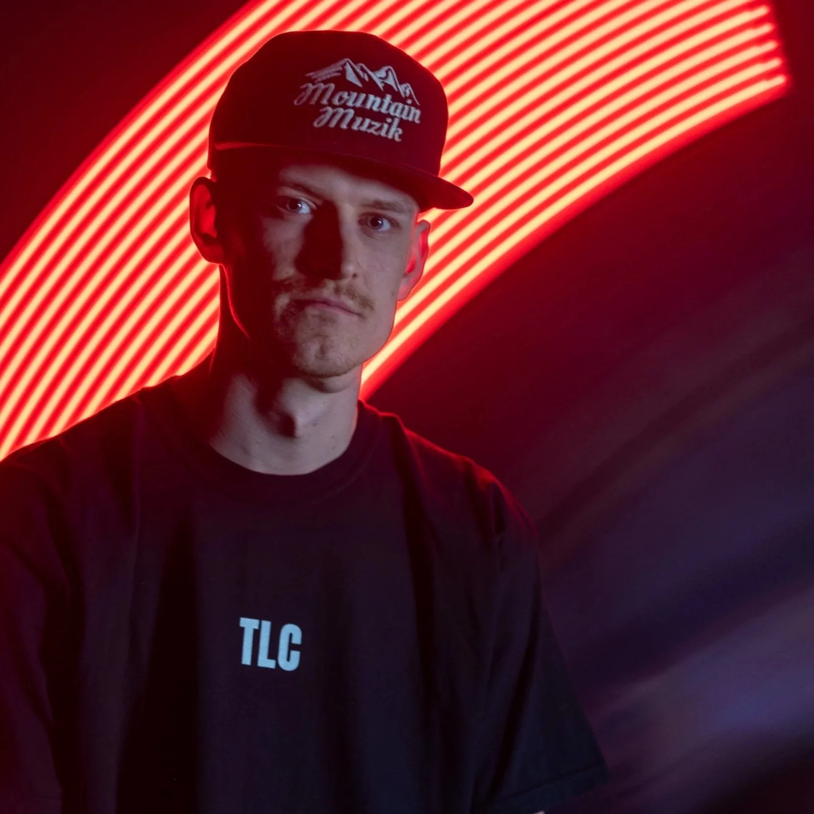 TR. wearing a black shirt and a black baseball cap with the words 'Mountain Muzik' on it, standing in front of a backdrop of vibrant red and orange neon lights.