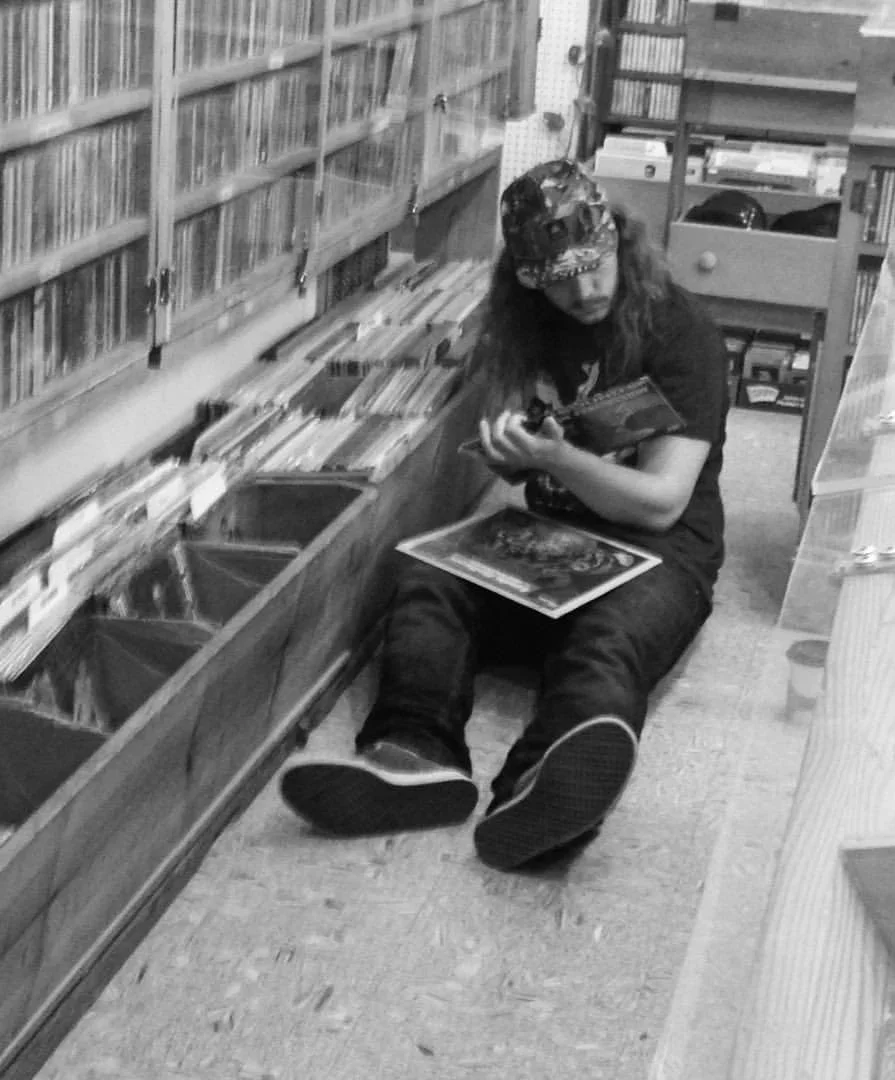 Symon sitting on the floor of a record store aisle, holding a record, with another one resting on his lap. He is surrounded by shelves filled with vinyl records.