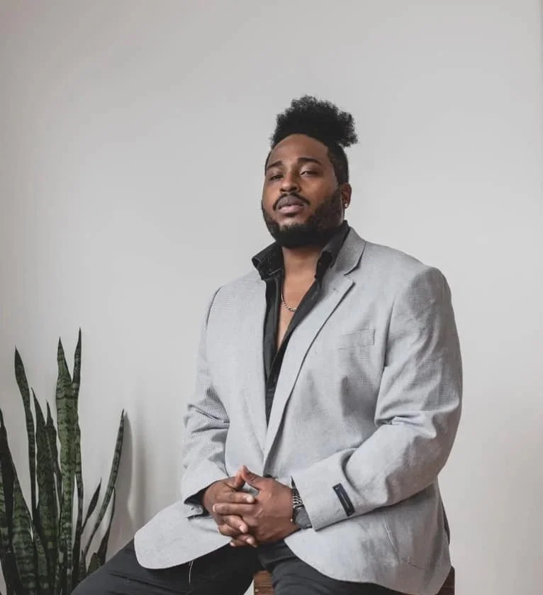 Atryp sitting against a plain wall, wearing a light gray blazer over a black shirt, with a silver watch and chain, posing confidently.