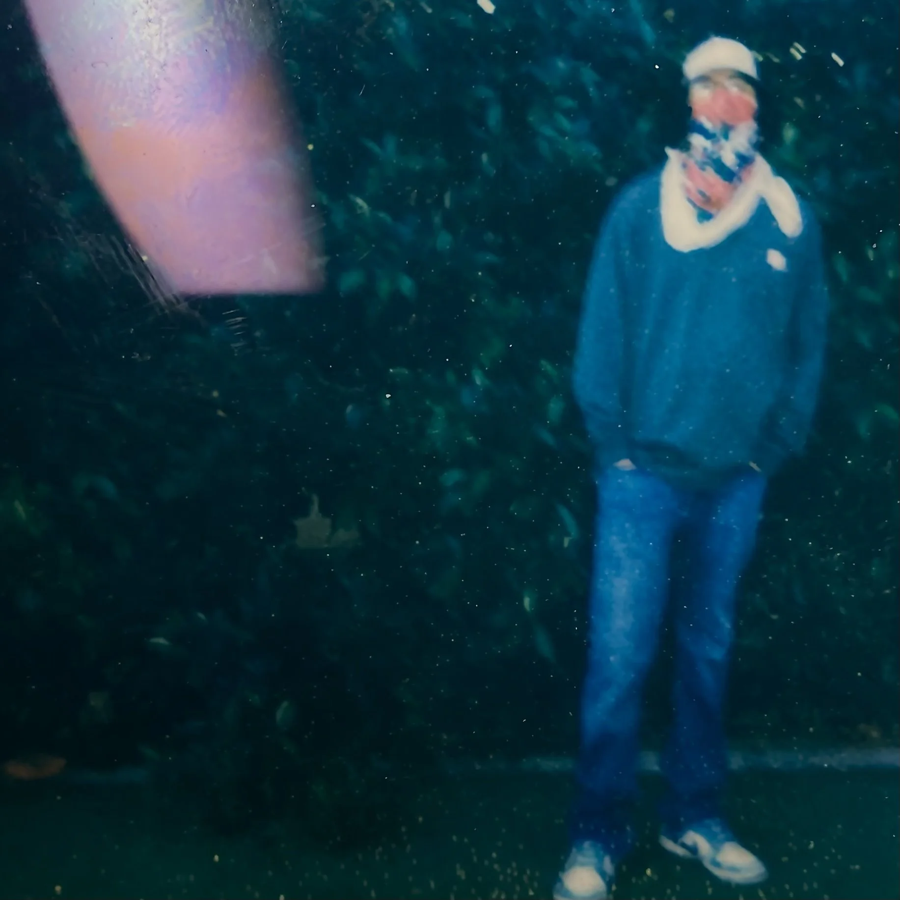 That Kid Nova wearing glasses, a baseball cap, and a colorful bandana standing outside in a nighttime setting with greenery background.
