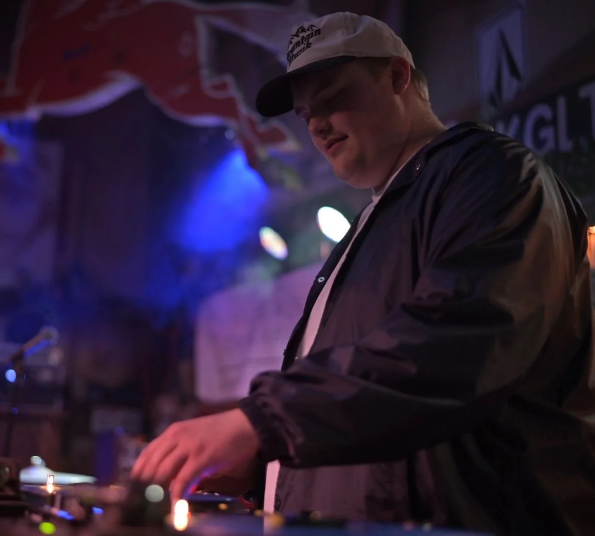 Mellow Mike DJing at a bar or club with colorful stage lighting and a casual style, wearing a baseball cap and a jacket.