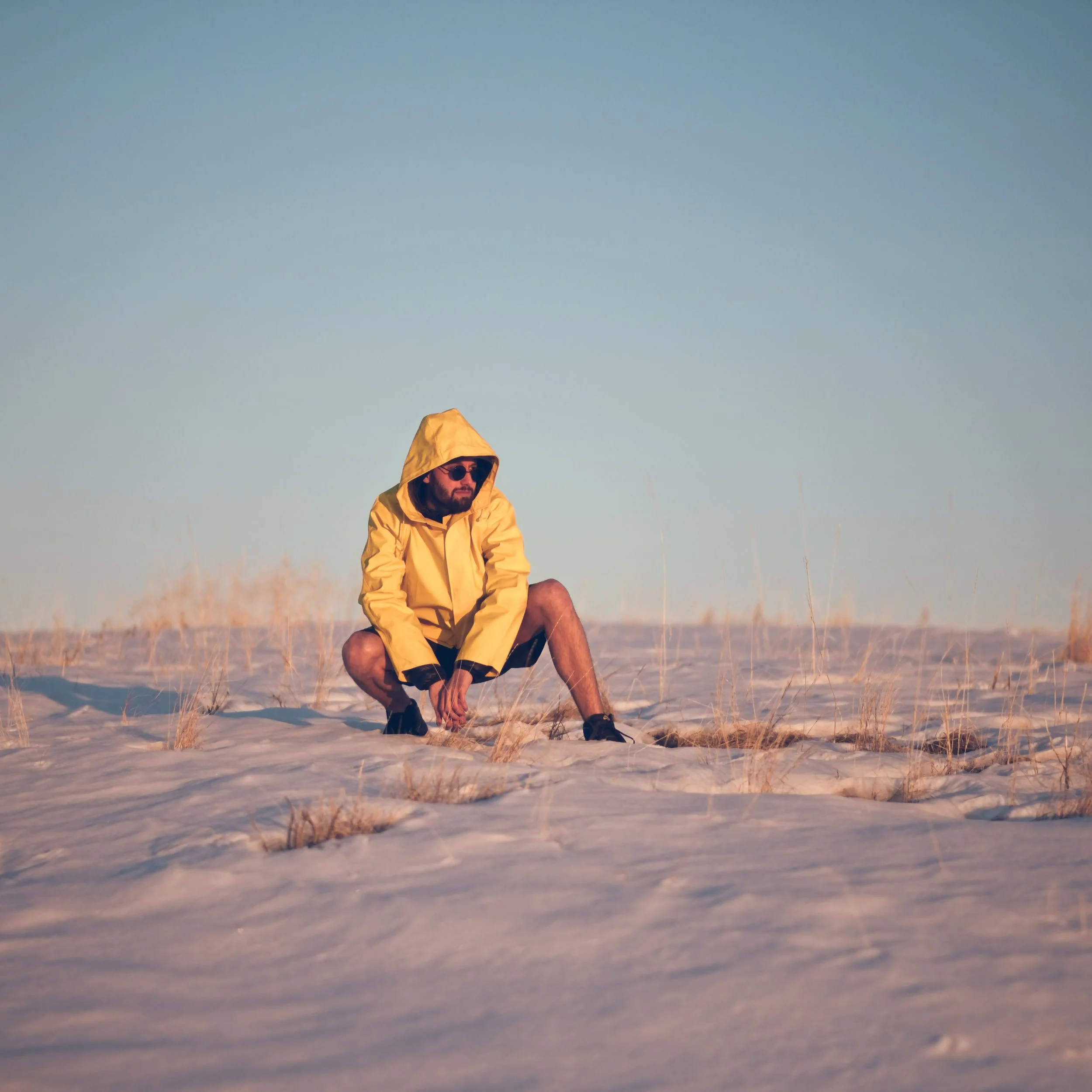 Twelve Clouds wearing a yellow jacket and sunglasses crouching in a sandy field with dry grass, under a clear blue sky.