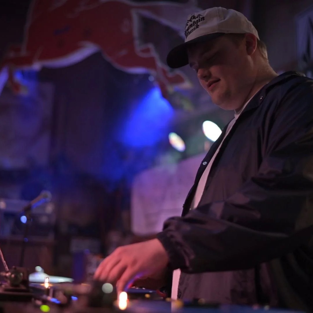 Mellow Mike wearing a baseball cap and a navy jacket is DJing at a dimly lit venue with blue lights and a red sign behind him on the wall.
