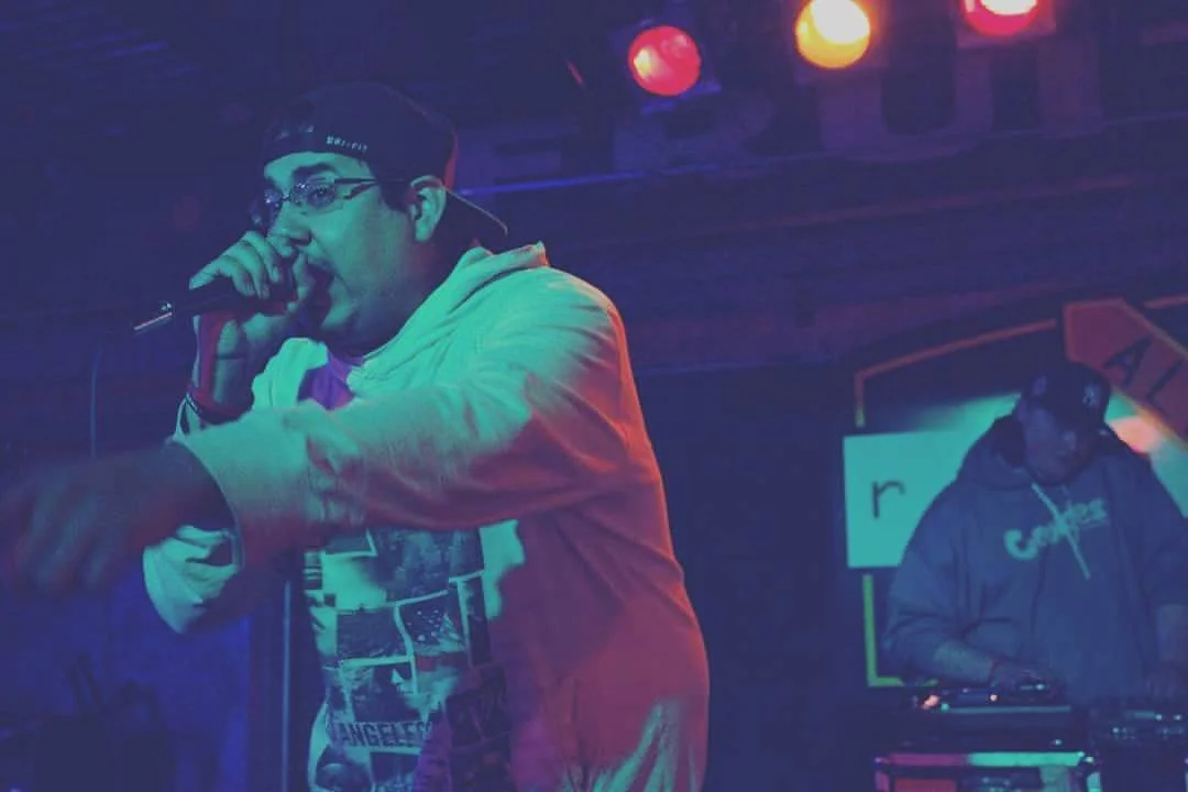 Image of C. Boy wearing glasses and a cap, holding a microphone, and performing on stage with colorful lighting, with Mellow Mike Djing in the background.