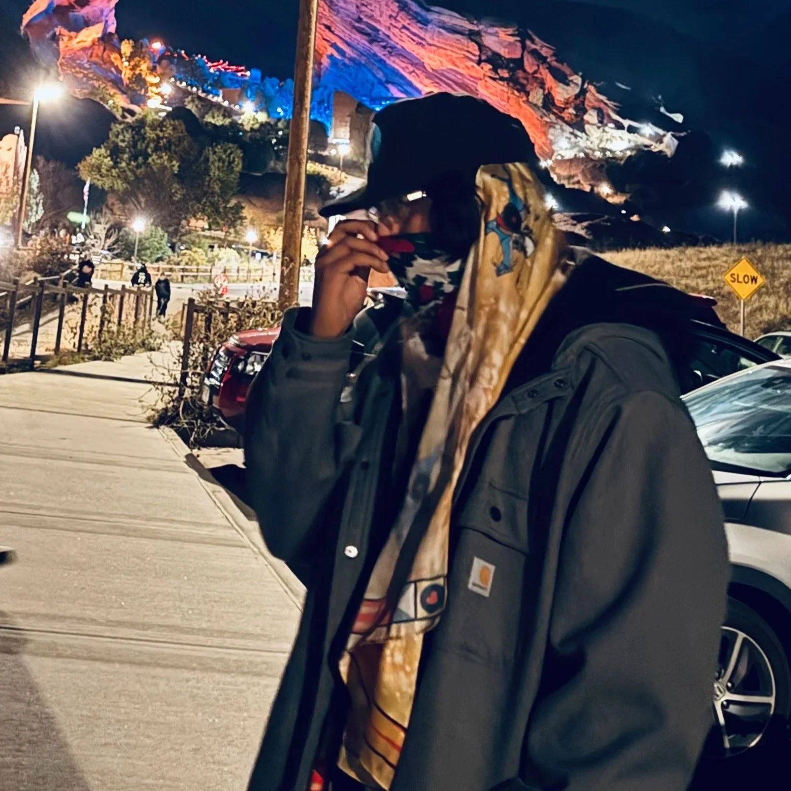 That Kid Nova, standing sideways wearing a jacket, long scarf, baseball hat, and glasses.  He's adjusting the bandana on his face, pulling it up with the Red Rocks Amphitheater in the background.