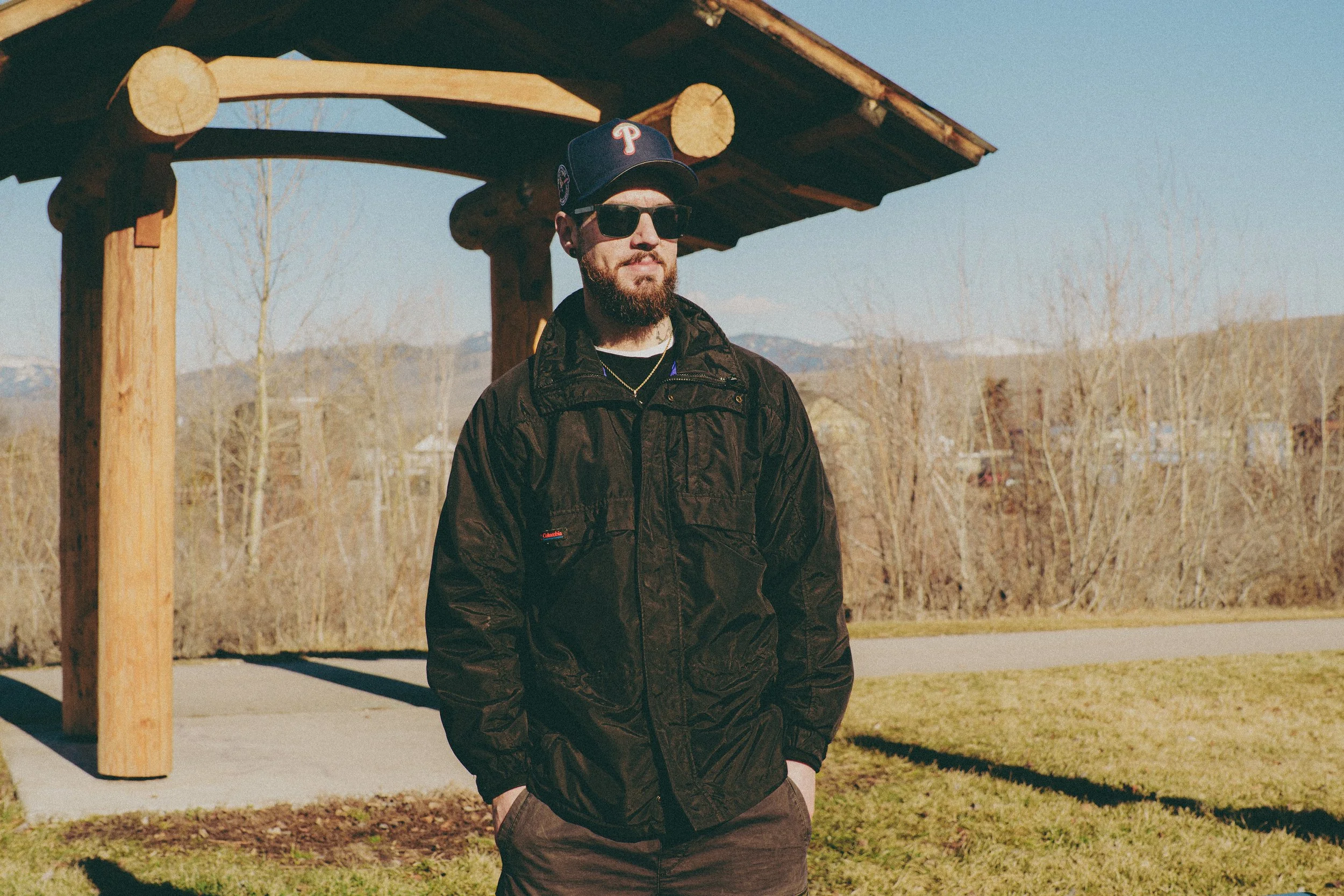 Yung Signal with a beard, wearing sunglasses, a black baseball cap with a red 'P', and a black jacket, stands outdoors under a wooden structure with trees and mountains in the background.