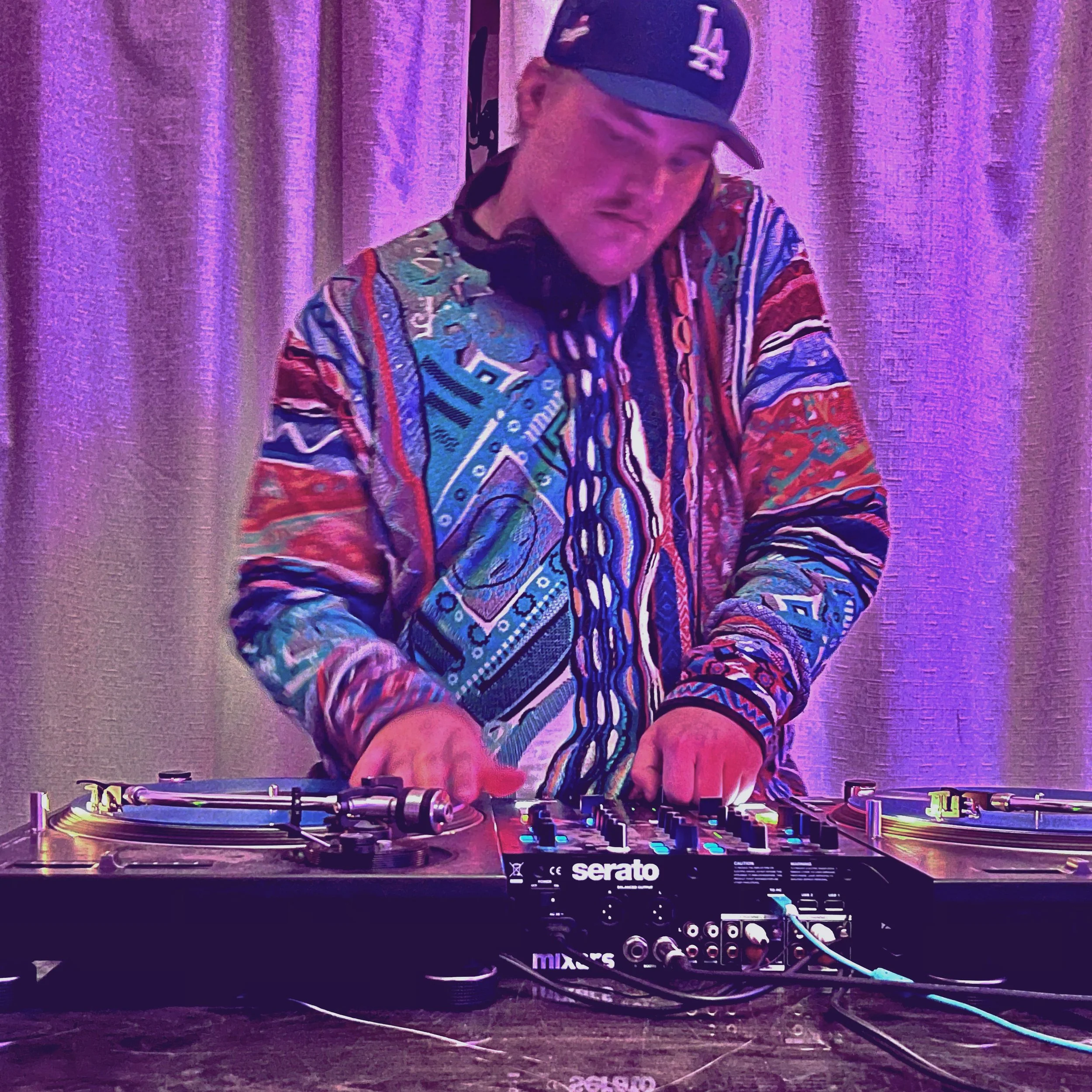 Mellow Mike DJing, wearing a colorful patterned shirt and a baseball cap with the LA Dodgers logo, standing behind turntables in a dimly lit room with purple lighting.