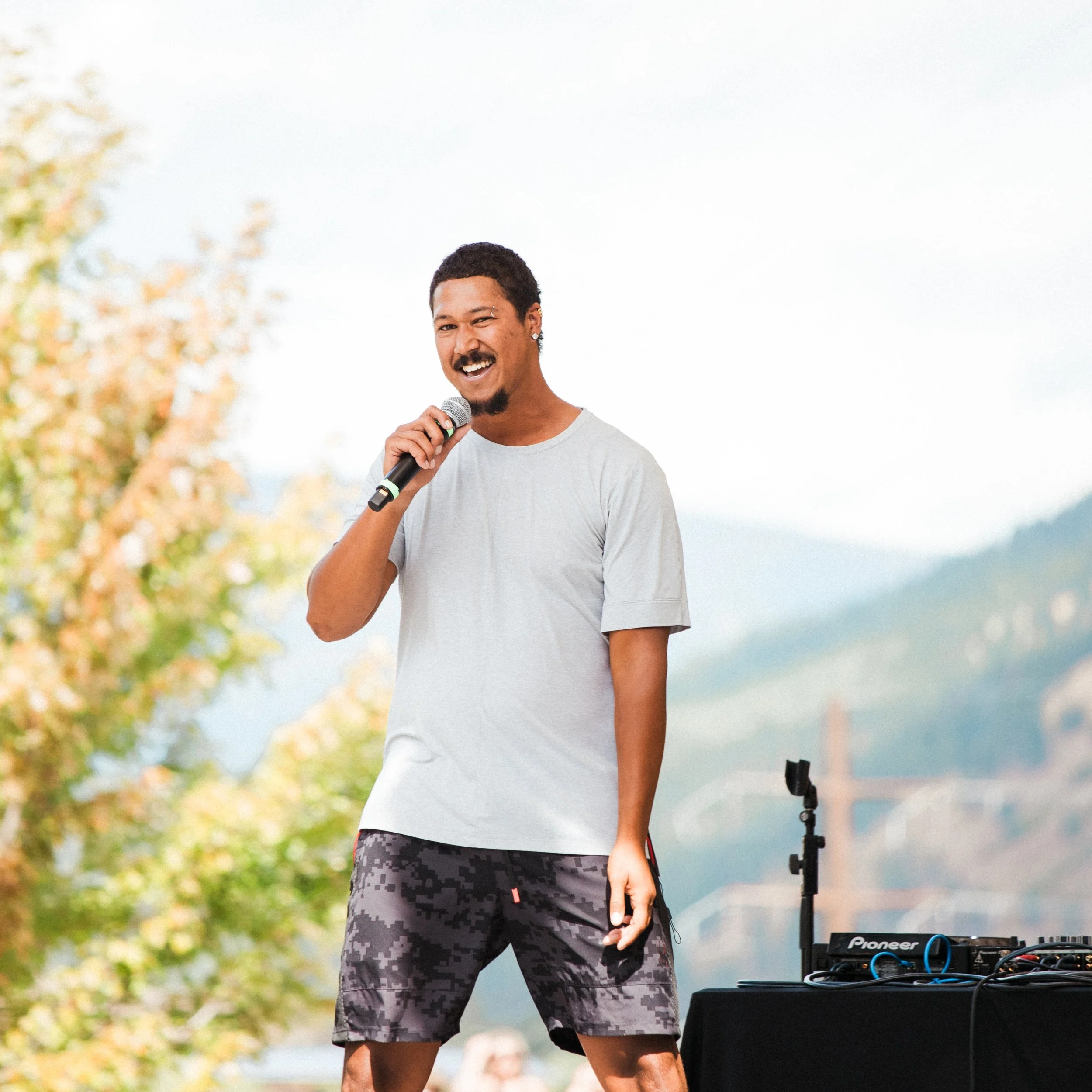R'Know holding a microphone, standing near DJ equipment outdoors, with trees and cloudy sky in the background.