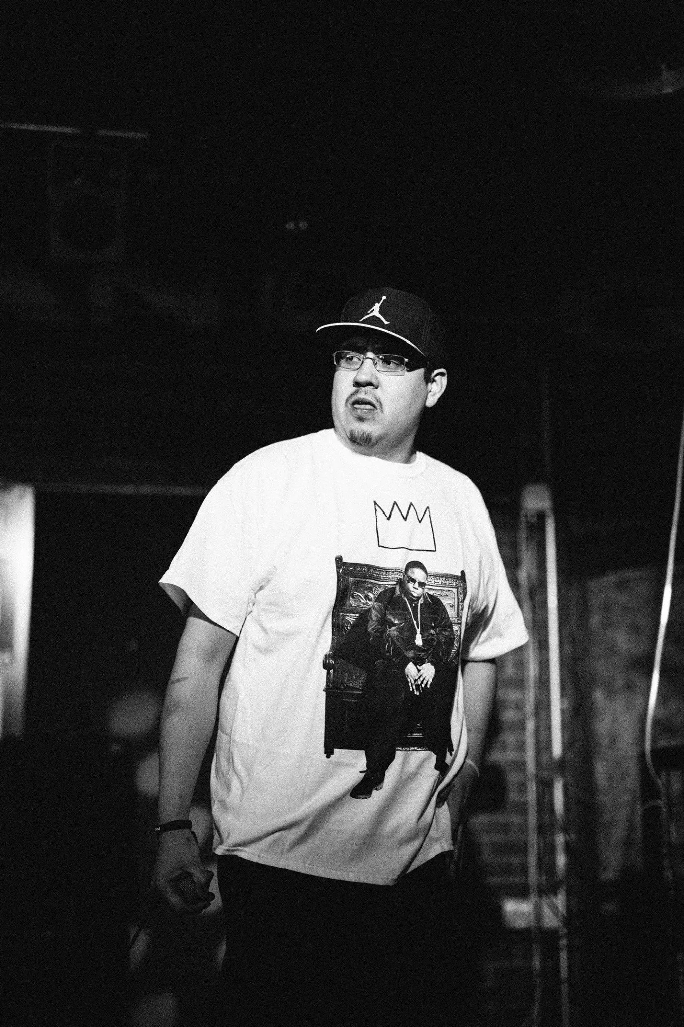 C. Boy wearing glasses, a black baseball cap with a Jumpman logo, and a graphic T-shirt, standing in a dark room and looking to his right.