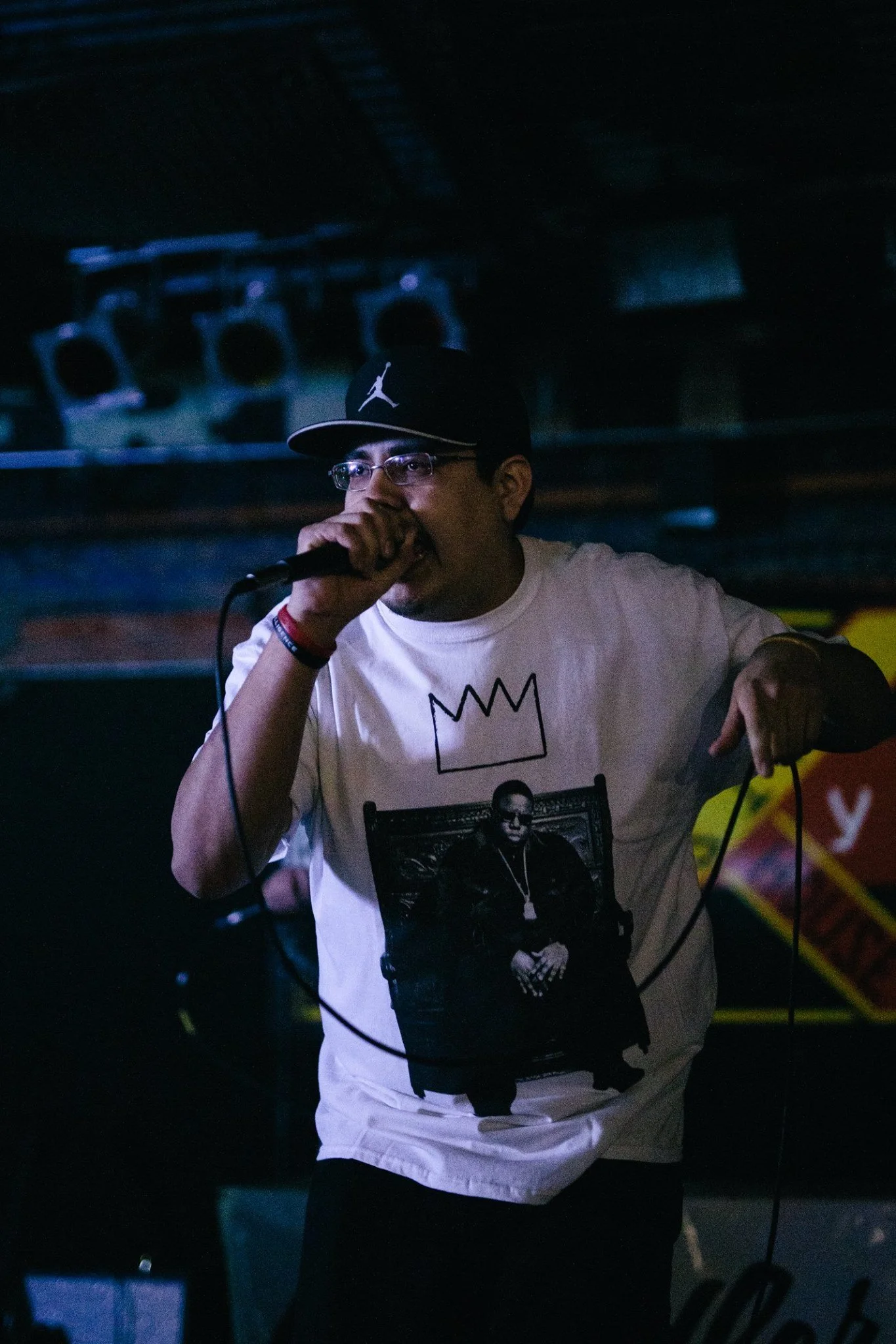 C. Boy wearing glasses, a black baseball cap with the Jordan logo, and a white T-shirt with a black and white graphic, holding a microphone and performing on stage in a dimly lit environment.