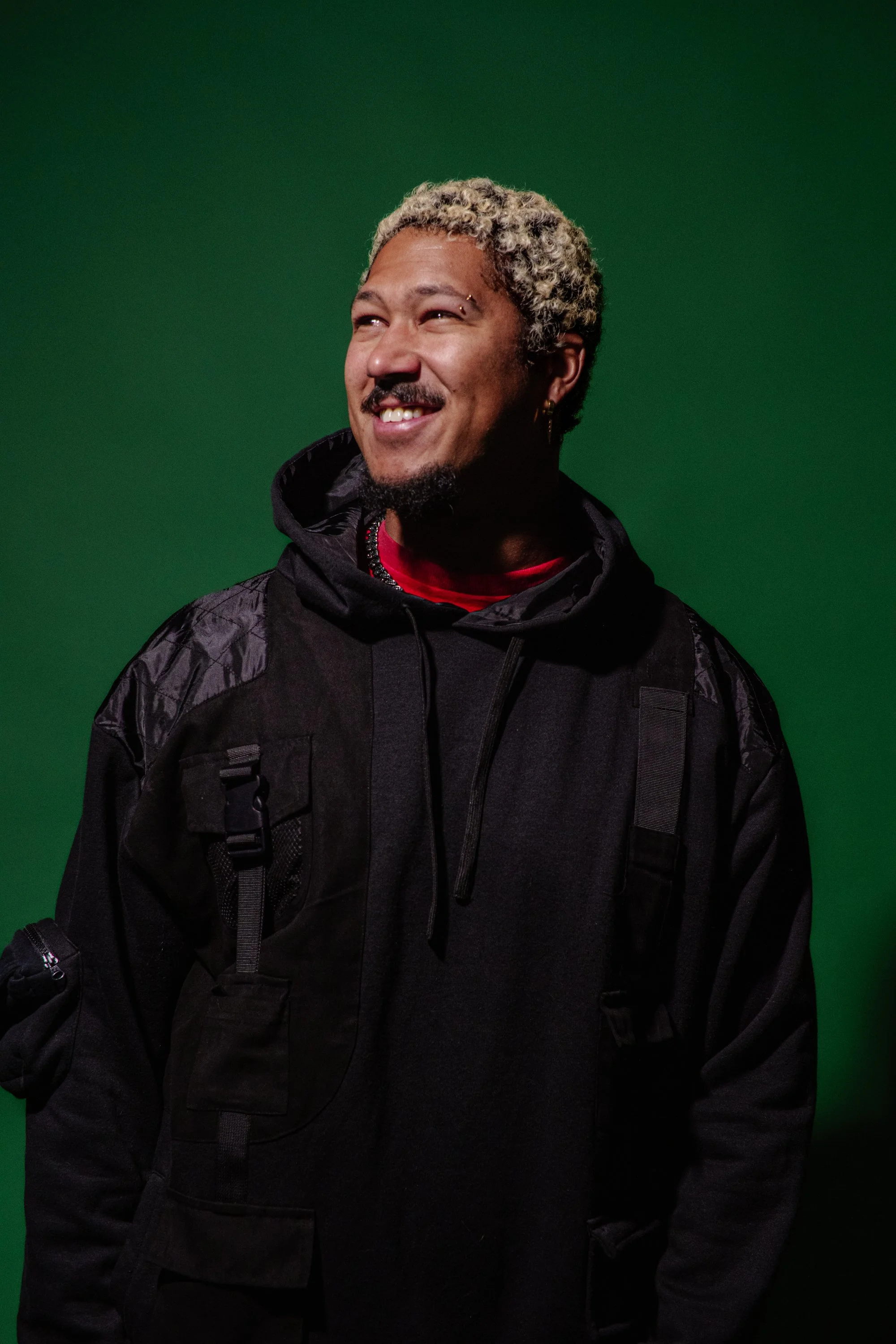 Photo of R'Know wearing a black jacket and a red shirt, standing against a green background.