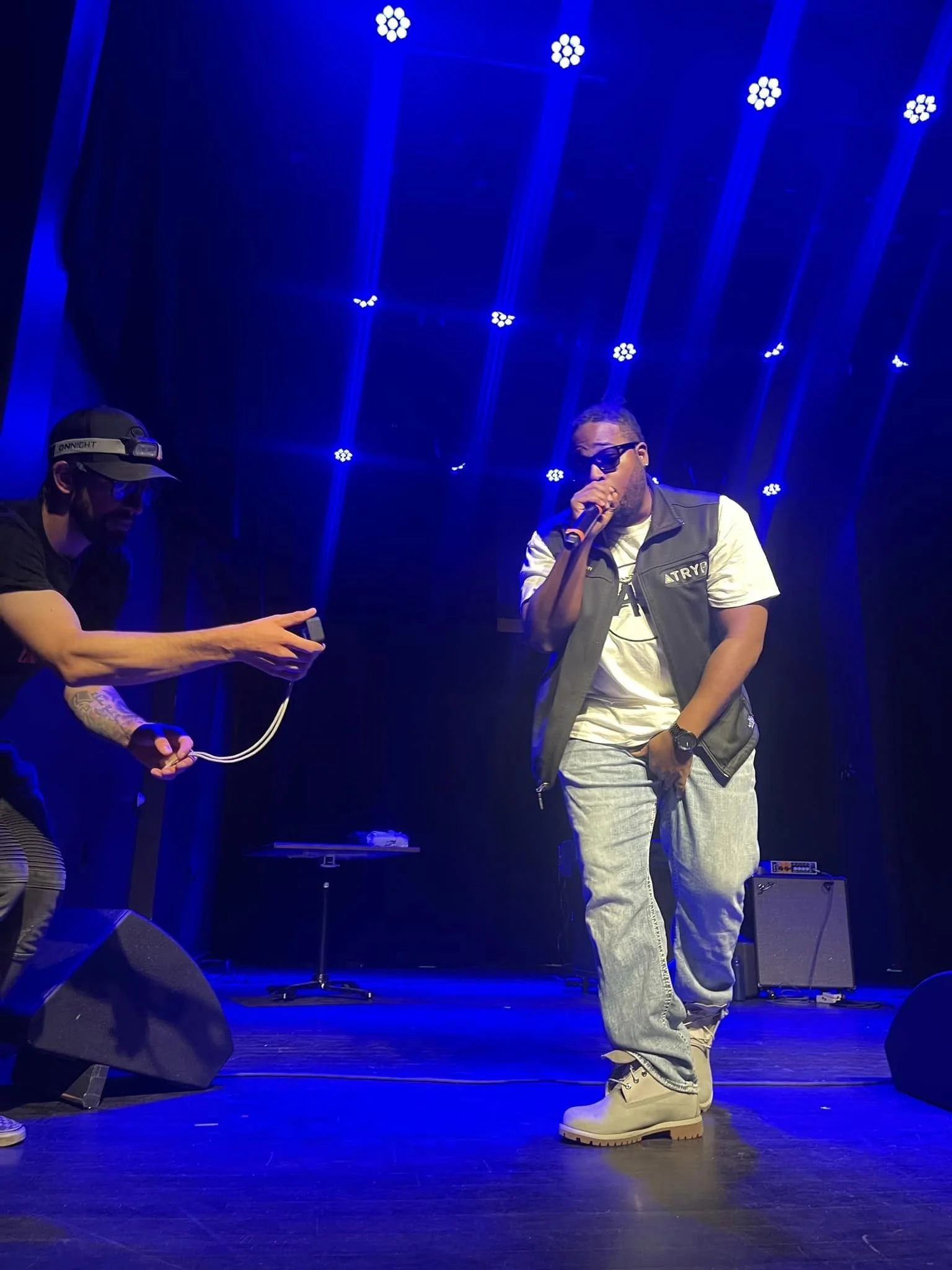 Atryp is performing with sunglasses, a gray vest, and baggy jeans performing on stage under blue lights, with a person on the left taking a photo or recording with a smartphone.